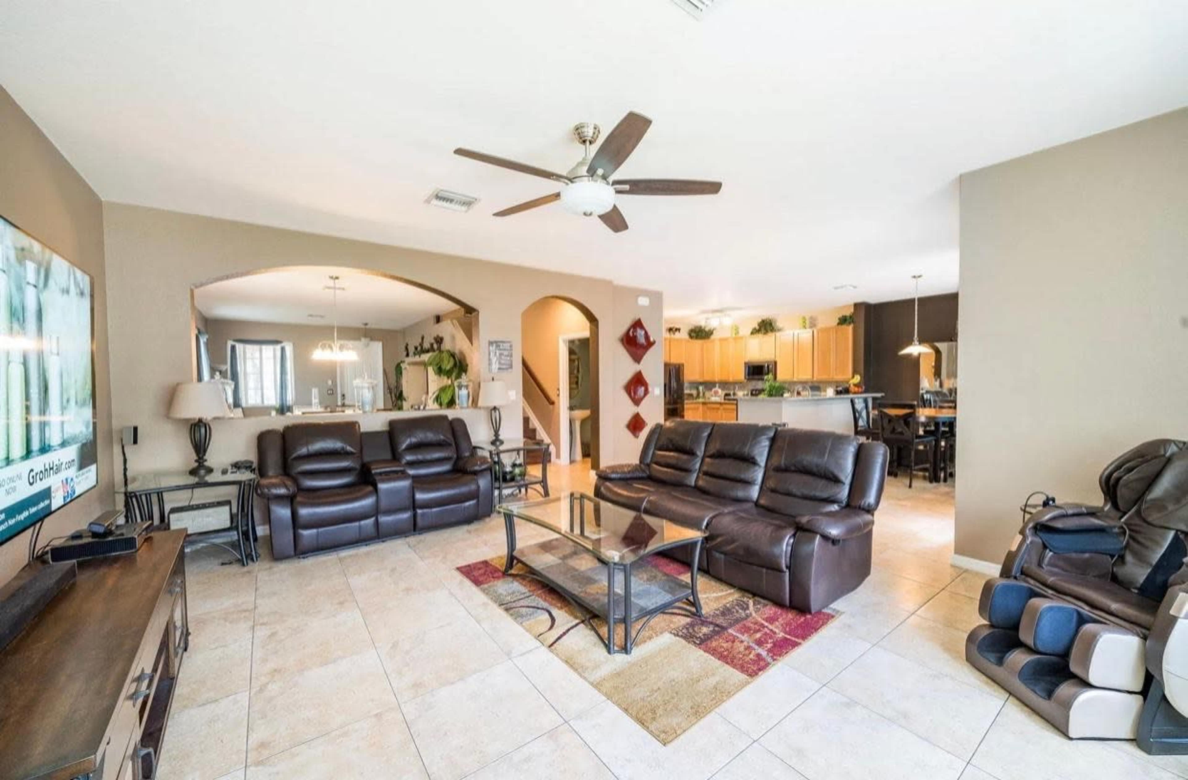 A spacious living room features dark leather sofas, a glass coffee table, and a TV, with an open view to a kitchen area in the background.