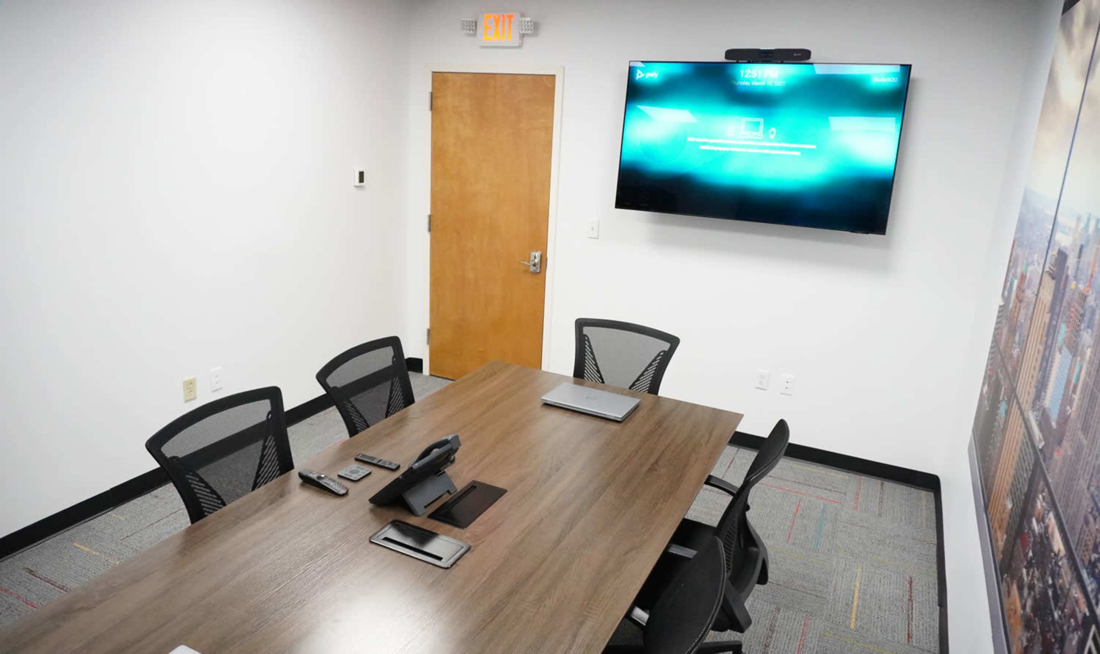 The image shows a modern conference room with a wooden table, several chairs arranged around it, and a large screen mounted on the wall displaying a digital interface.