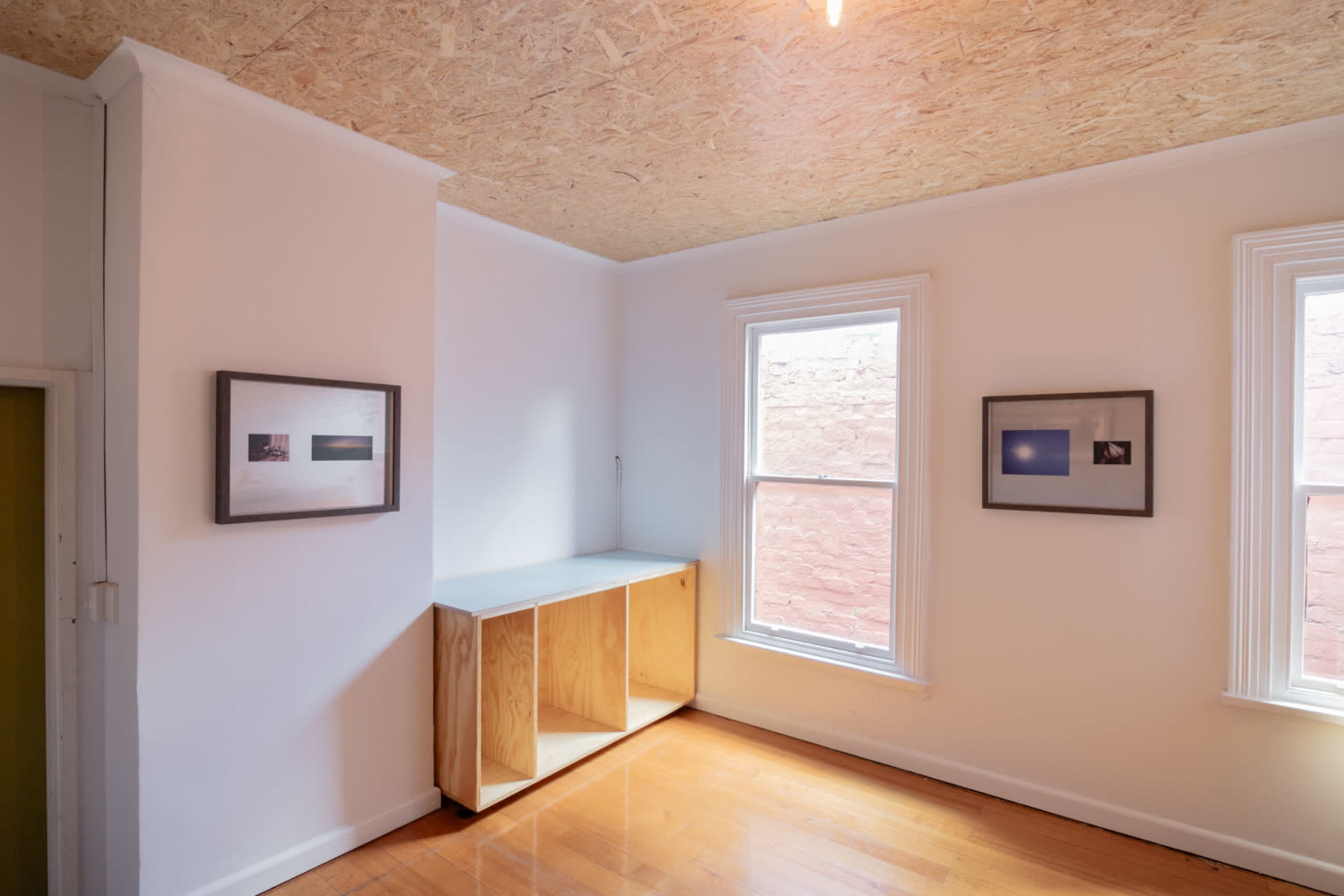 The image shows a minimalist room with white walls, two windows, and wooden flooring, featuring a simple wooden cabinet and framed photos on the walls.