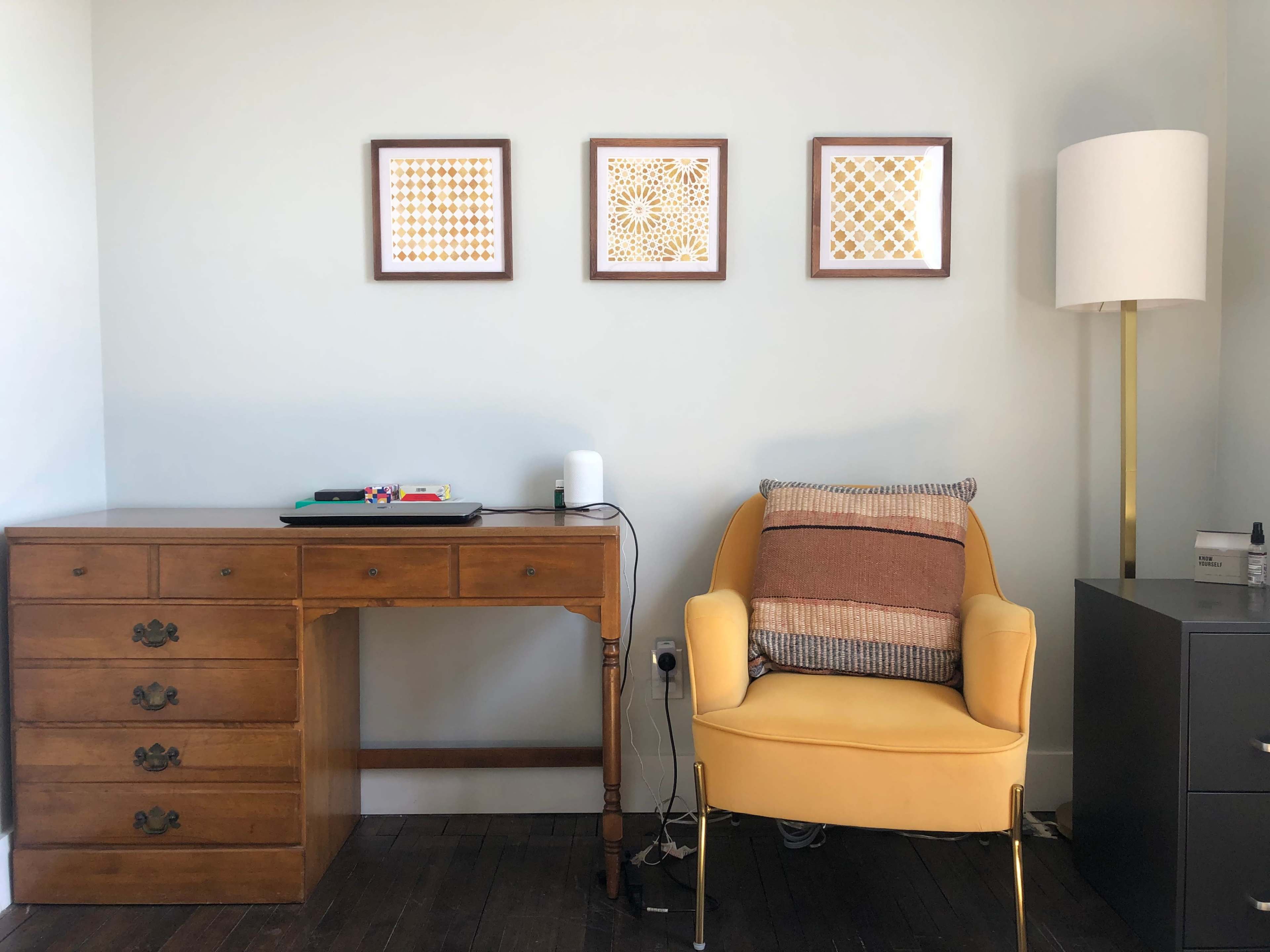The image depicts a room with a wooden desk, a yellow armchair, and three framed decorative wall pieces above the desk.