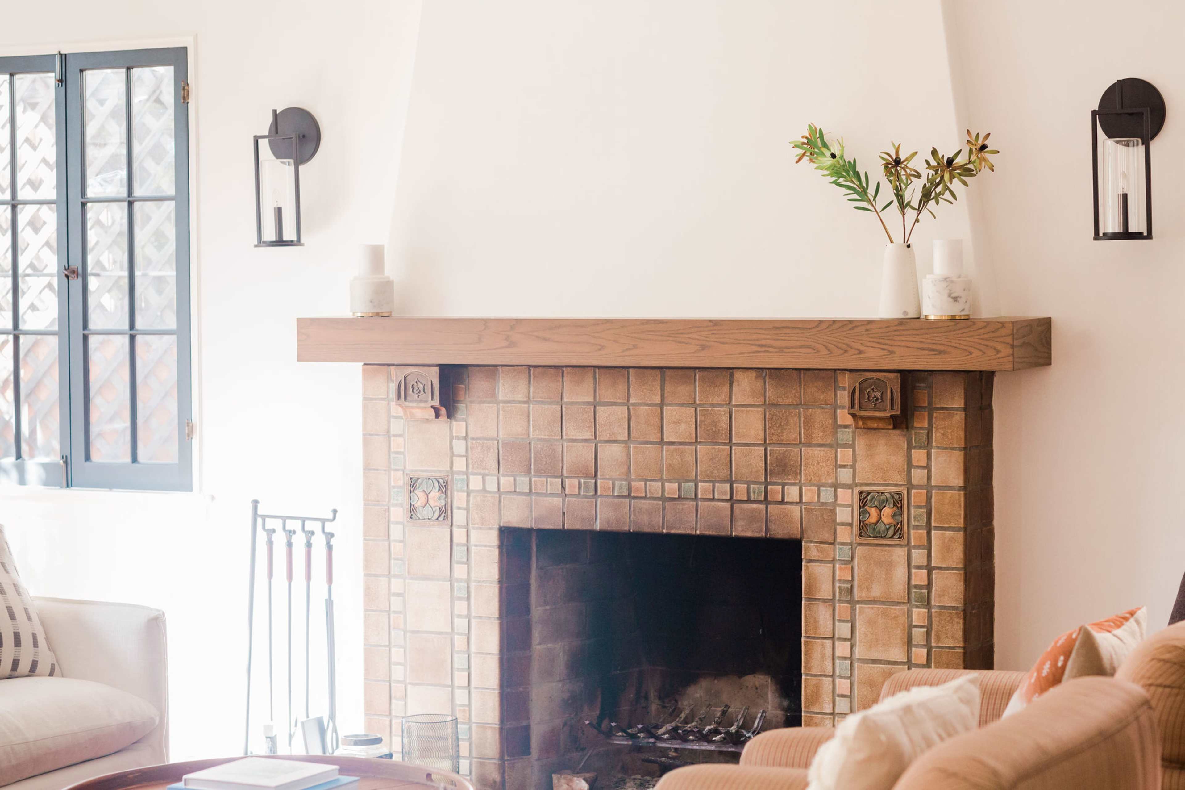 A living room with a tiled fireplace, wooden mantel, and decorative items on top, accompanied by a large window and comfortable seating.