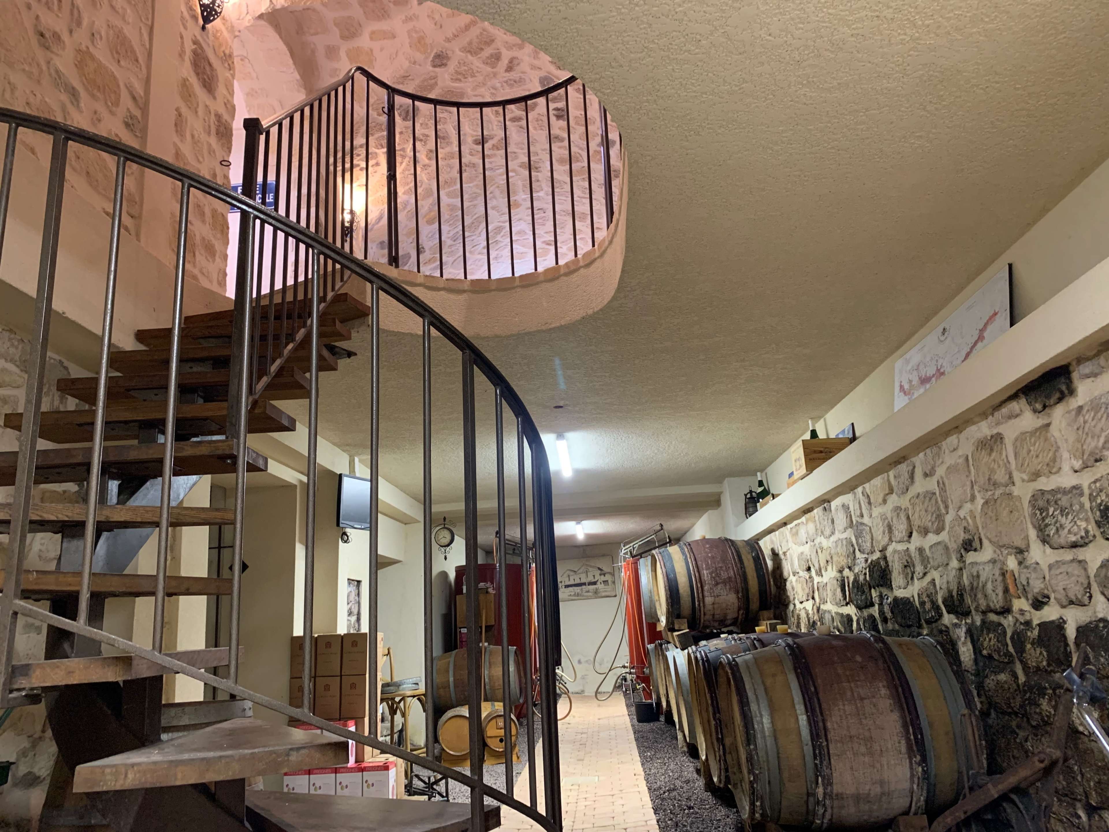 A spiral staircase leading to an upper level in a dimly lit wine cellar filled with wooden barrels and stone walls.