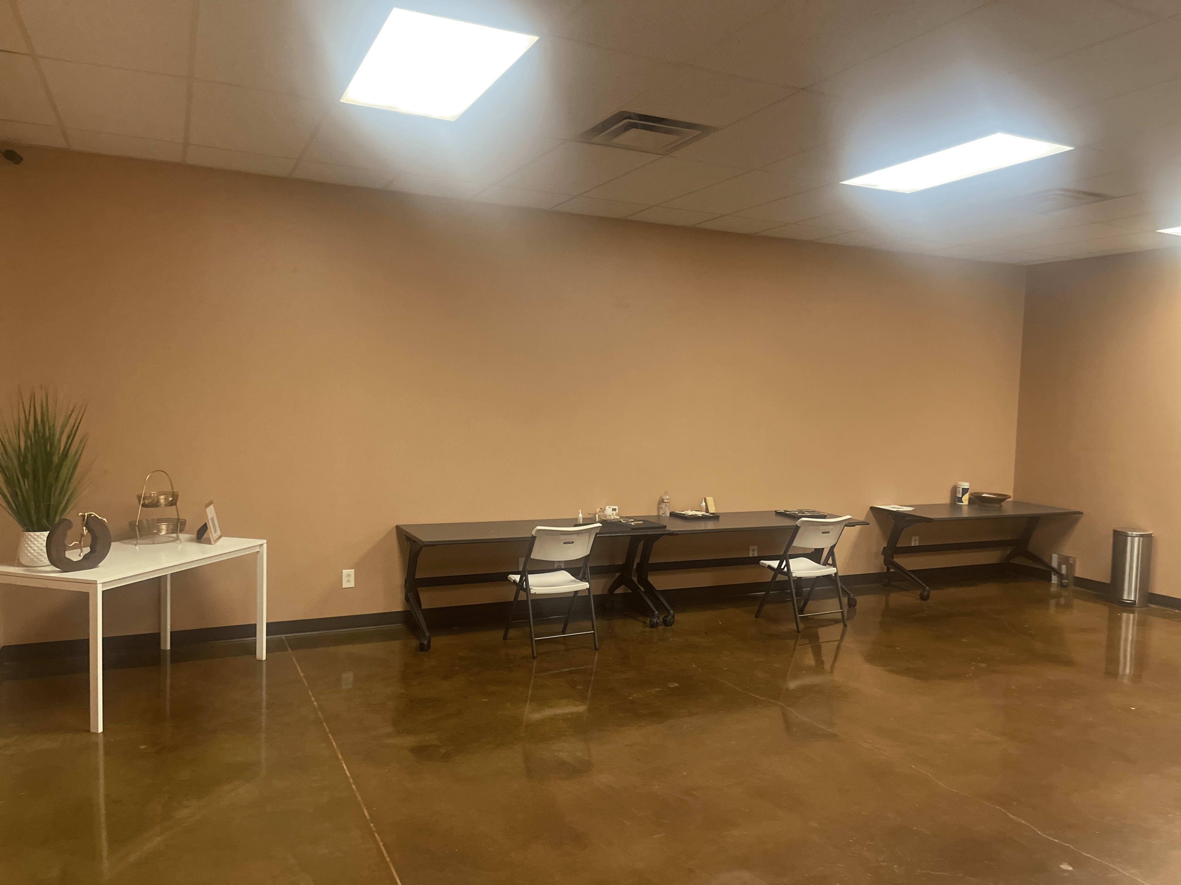 The image shows a room with a polished concrete floor, featuring several tables and chairs arranged against a plain wall, along with a decorative plant on a side table.