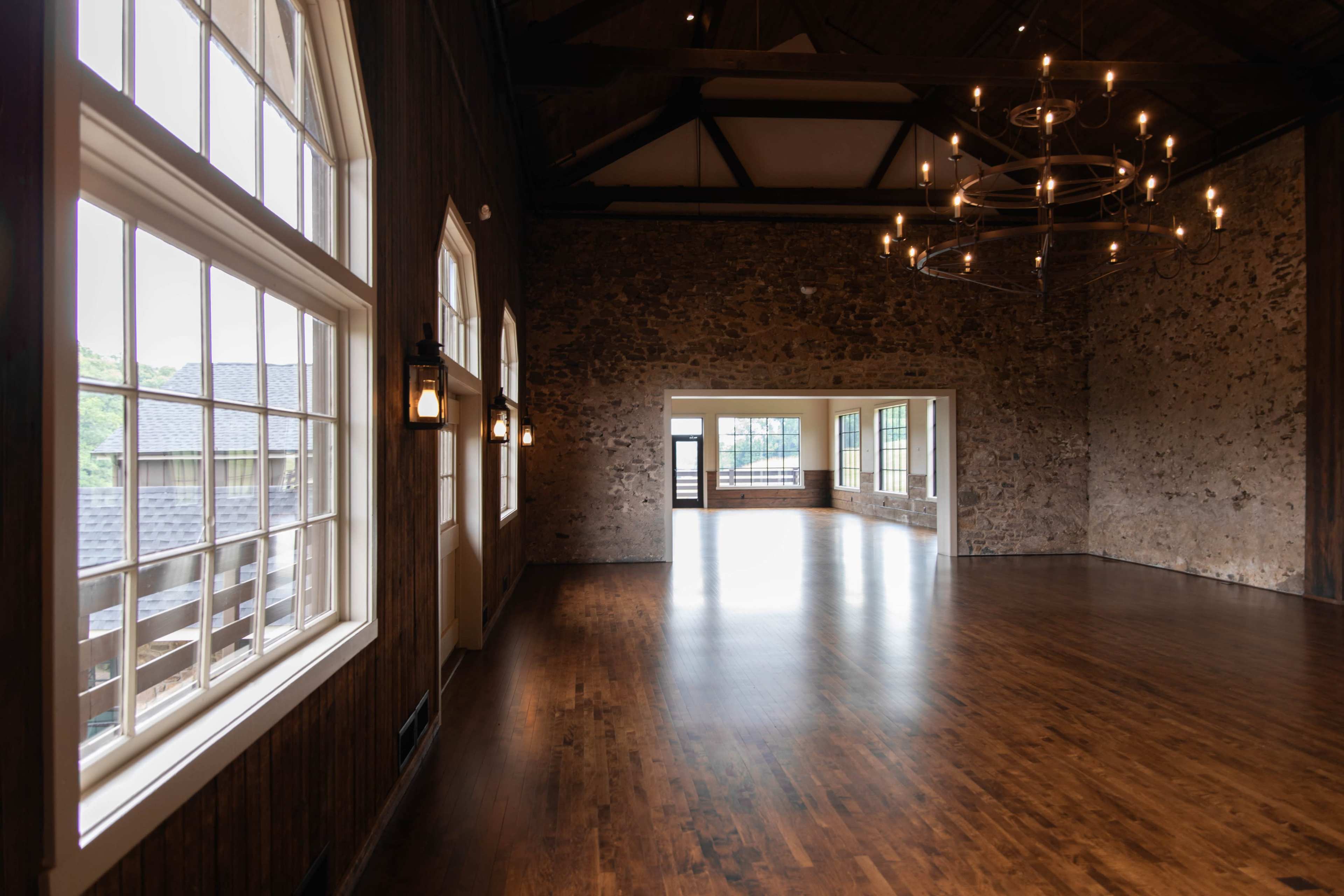 The image shows a spacious room with large windows and a wooden floor, featuring an illuminated chandelier and exposed stone walls.