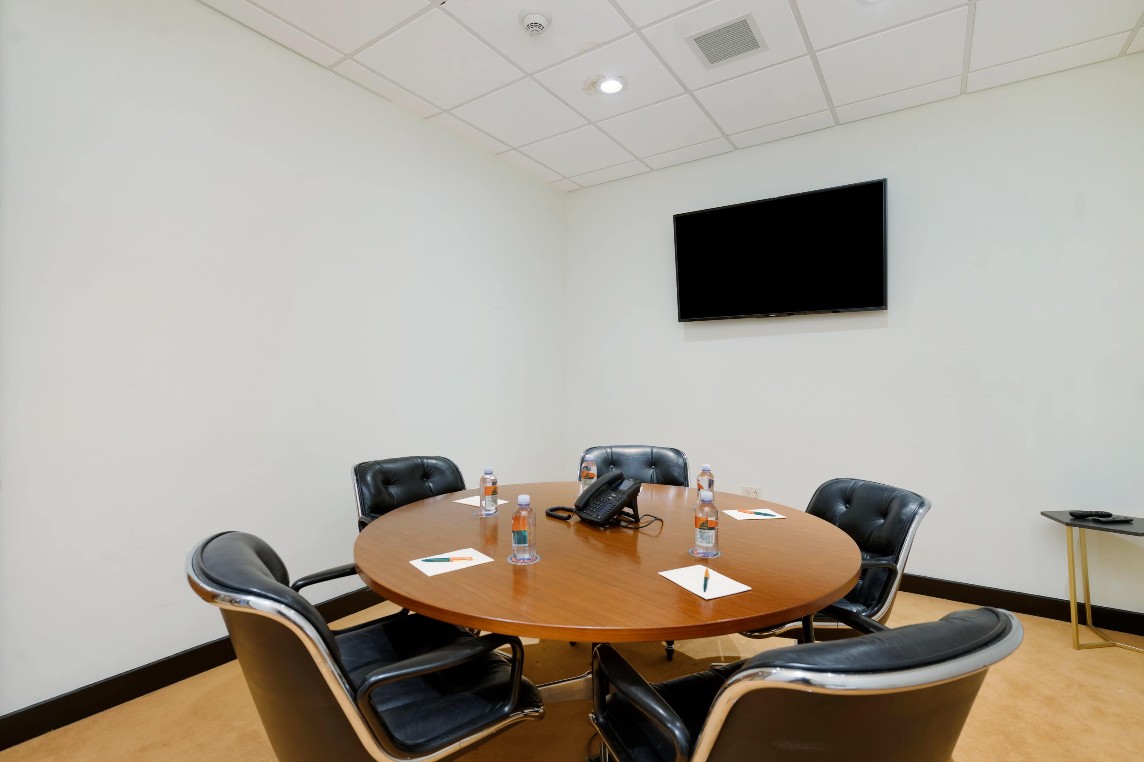 A round wooden conference table surrounded by five black leather chairs is set up in a meeting room, complete with a telephone and water bottles on the table, and a mounted television on the wall.