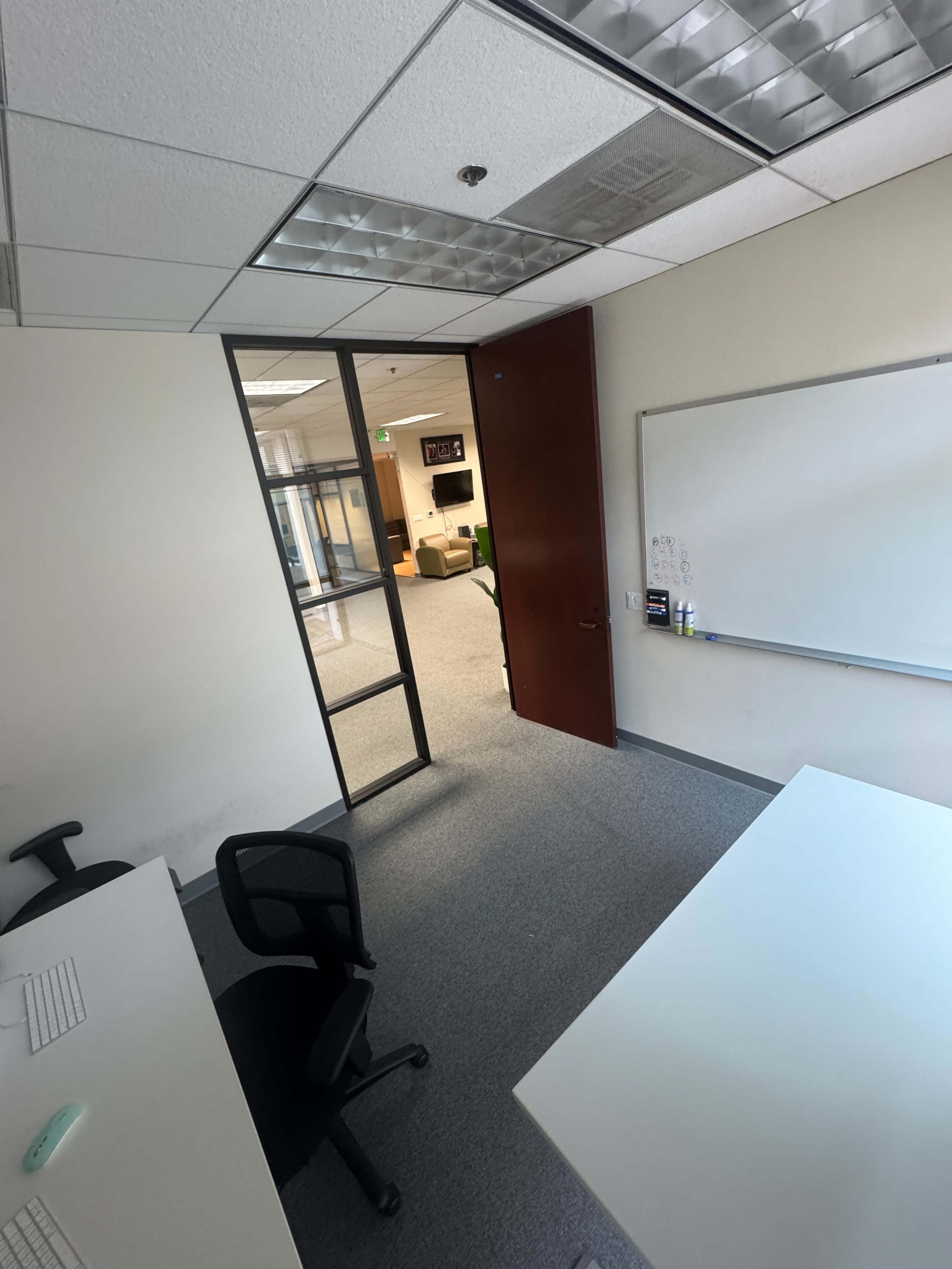 The image shows an office space with an open glass door leading to a corridor, two chairs positioned at desks, and a whiteboard on the wall.