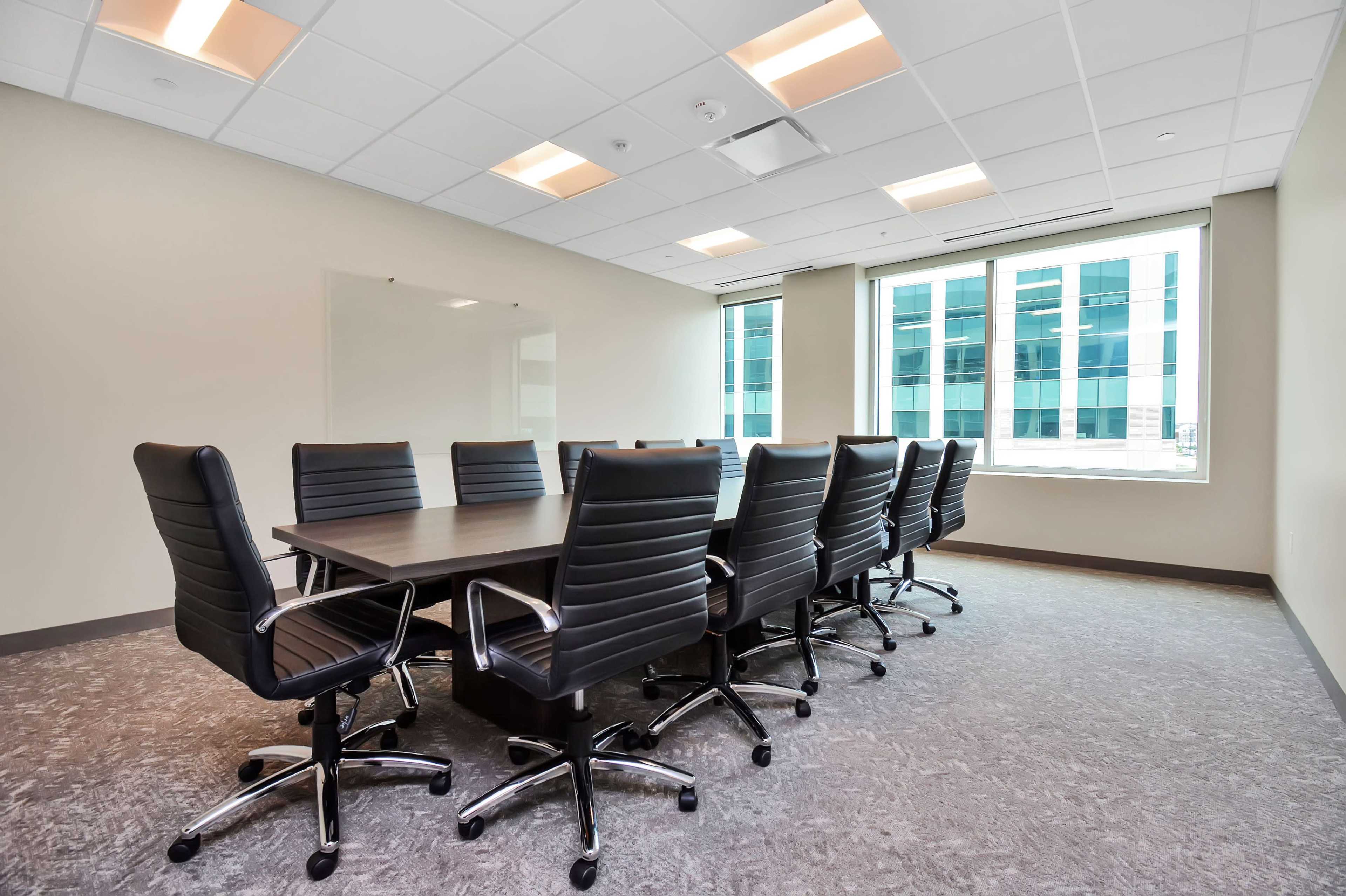 A large conference room features a rectangular table surrounded by eight black swivel chairs, with windows overlooking an urban landscape.