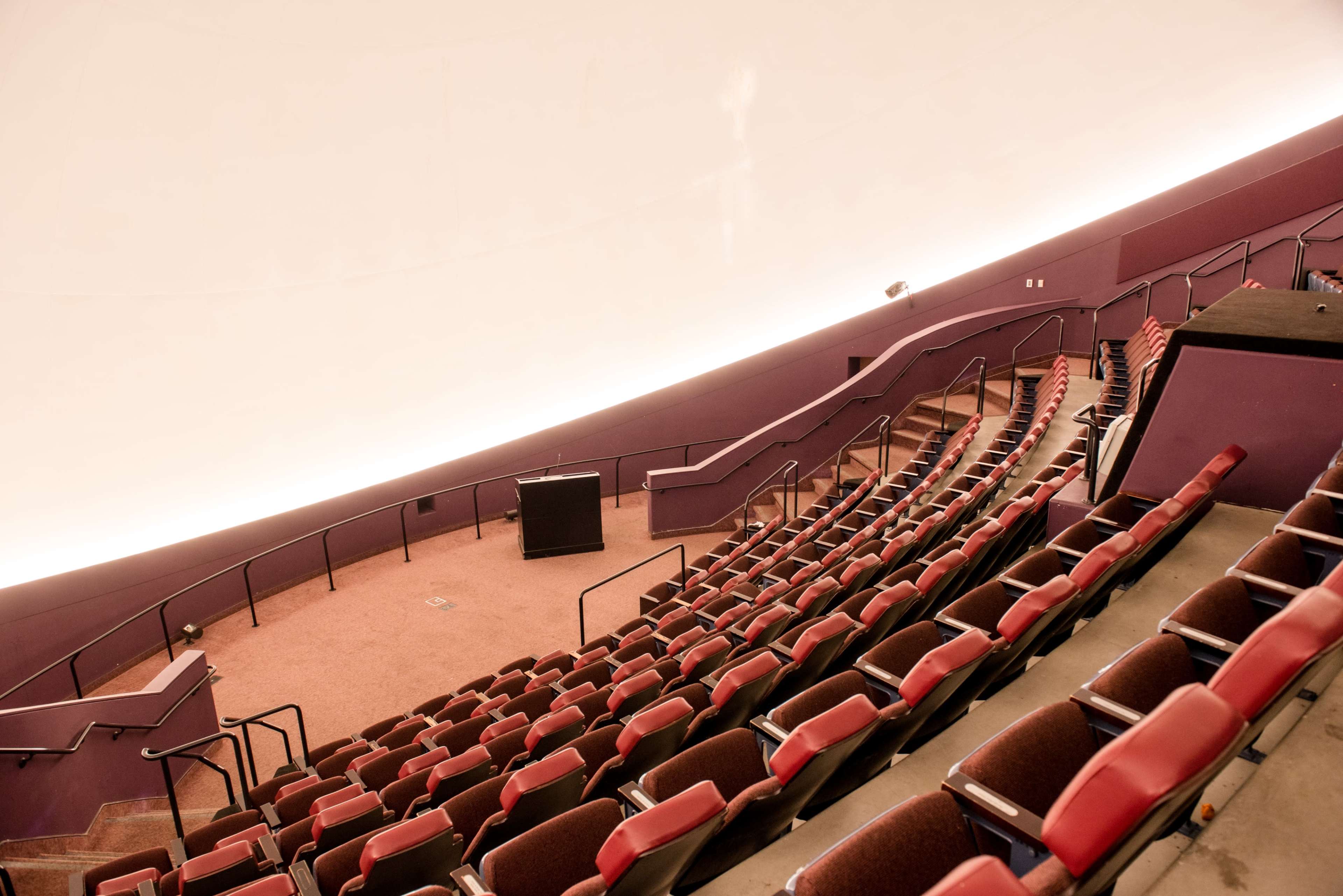The image shows a dimly lit auditorium with tiered seating and a large curved ceiling.