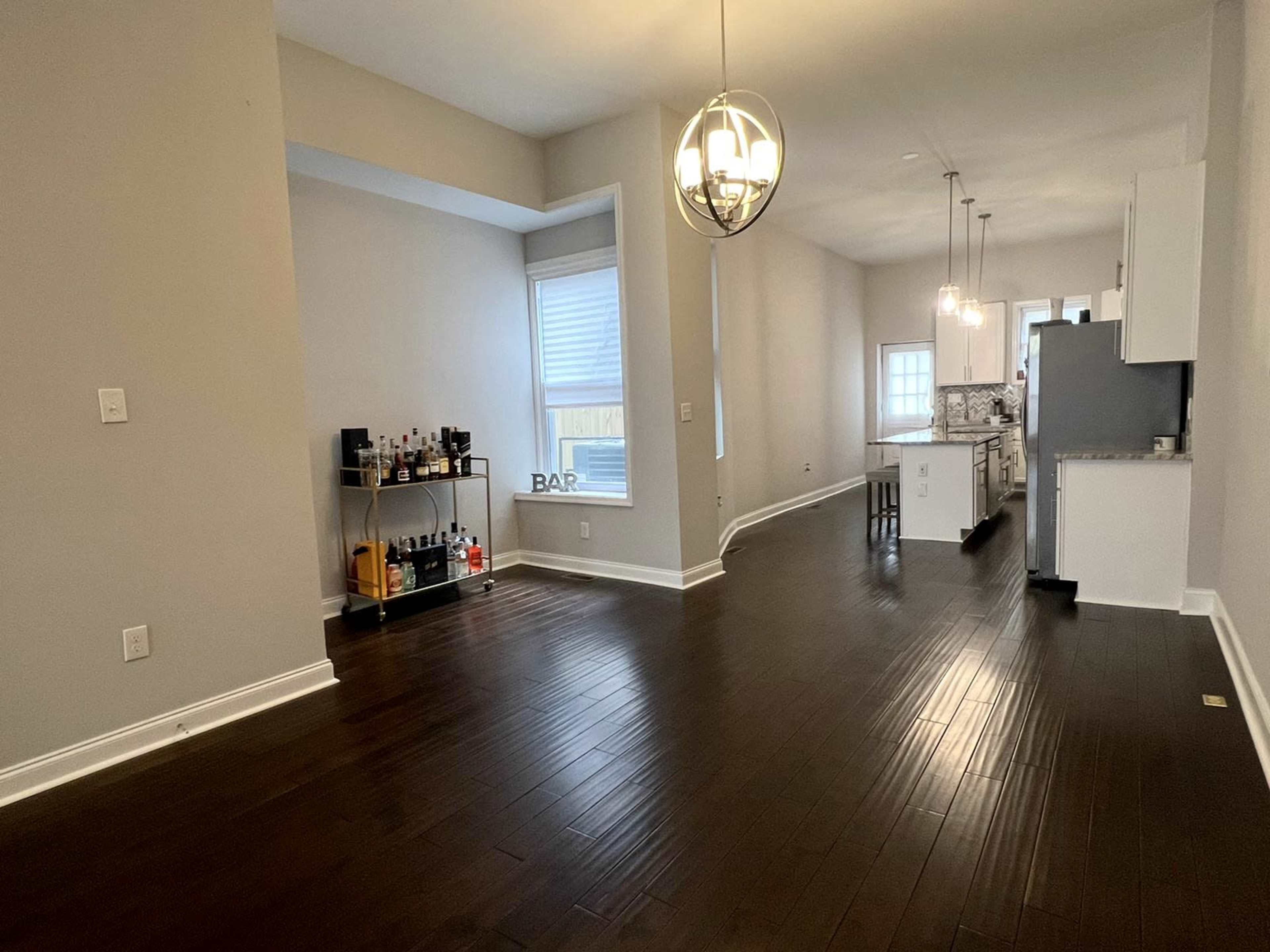 A spacious, bright room with dark hardwood floors, featuring a bar cart on the left and an open kitchen area in the background.