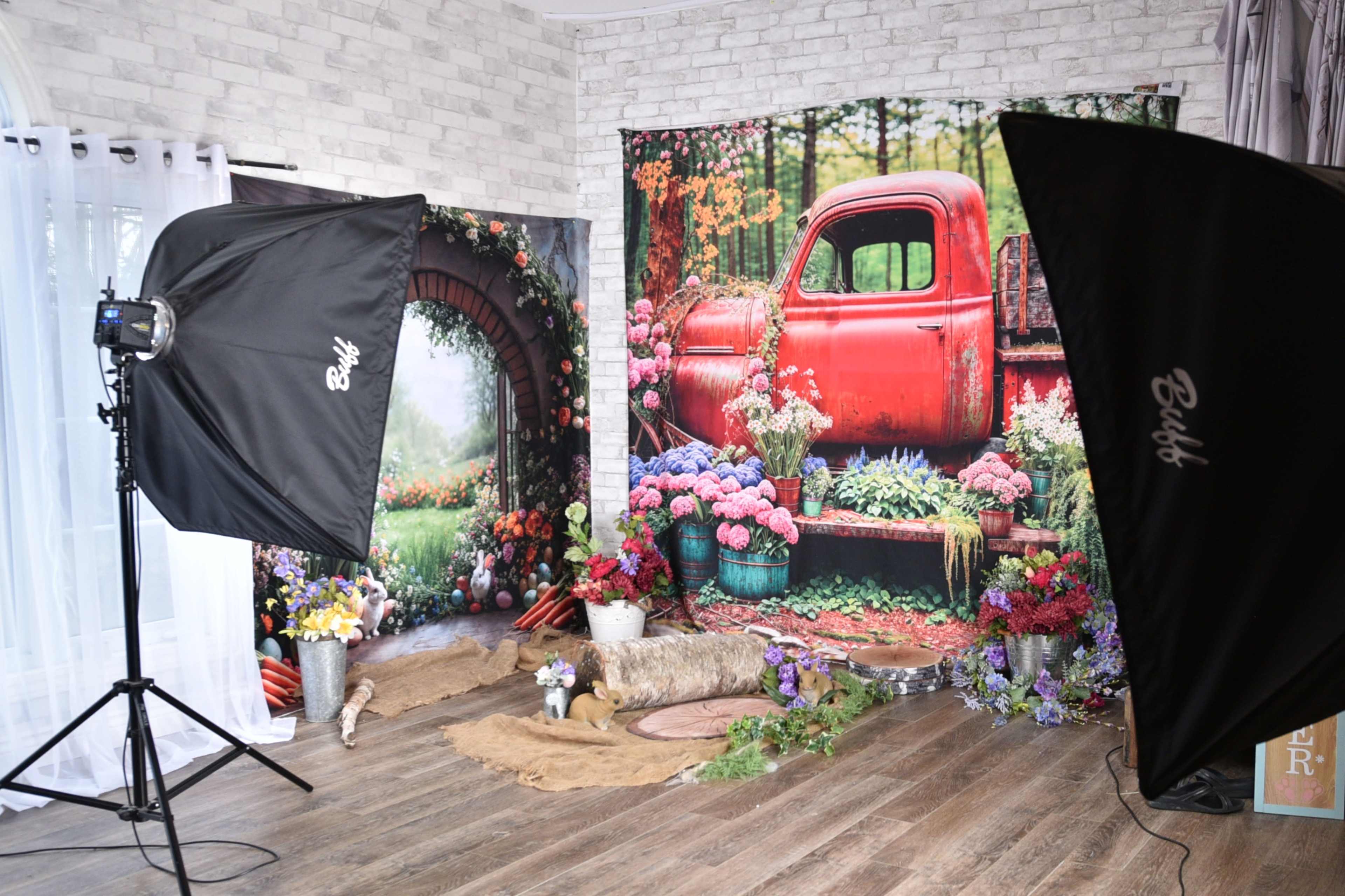 The image shows a photography studio setup featuring a backdrop of a red vintage truck amidst flowers and greenery, flanked by two lighting softboxes.