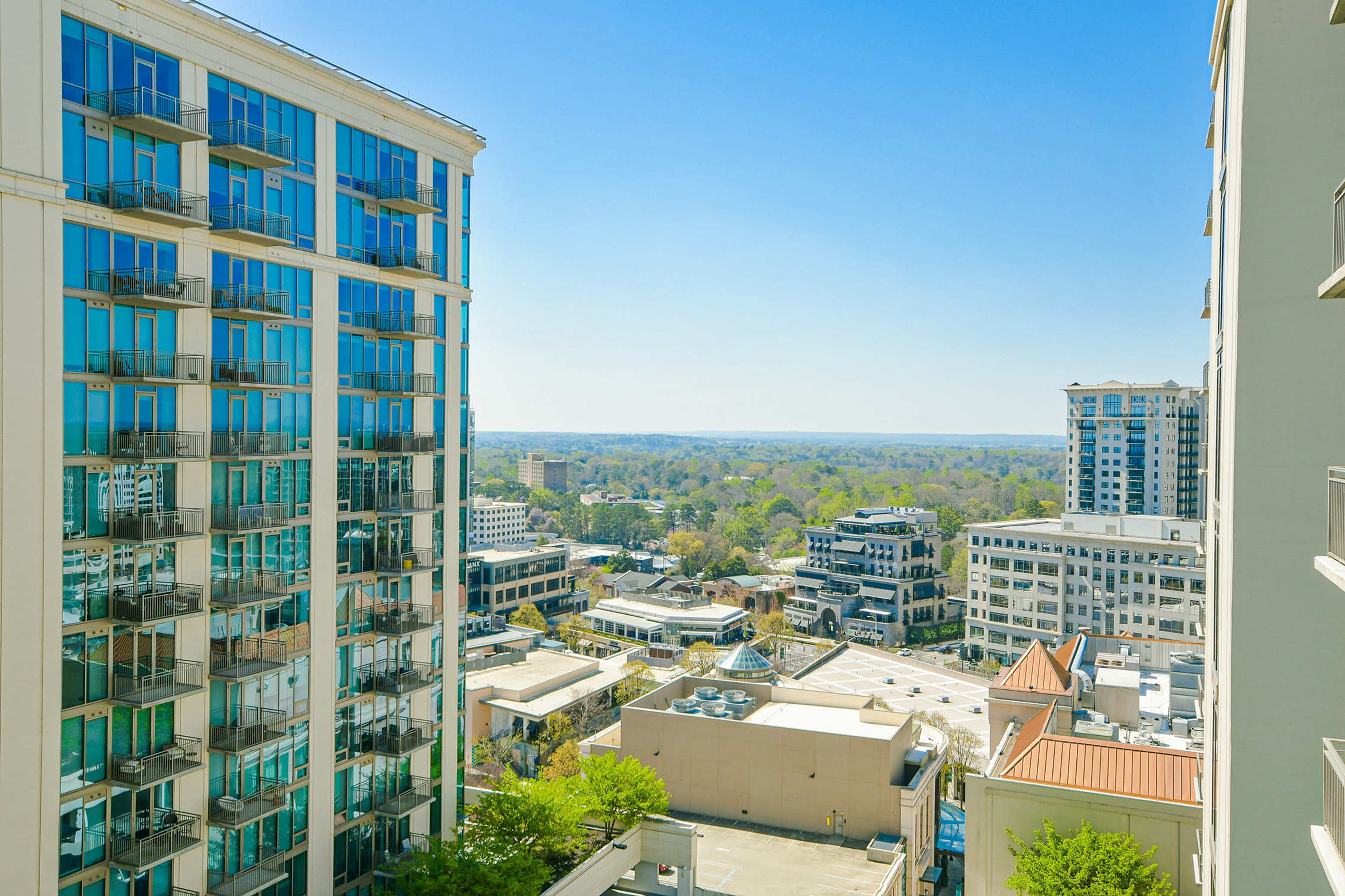 A panoramic view of a city skyline with modern buildings and greenery in the foreground under a clear blue sky.
