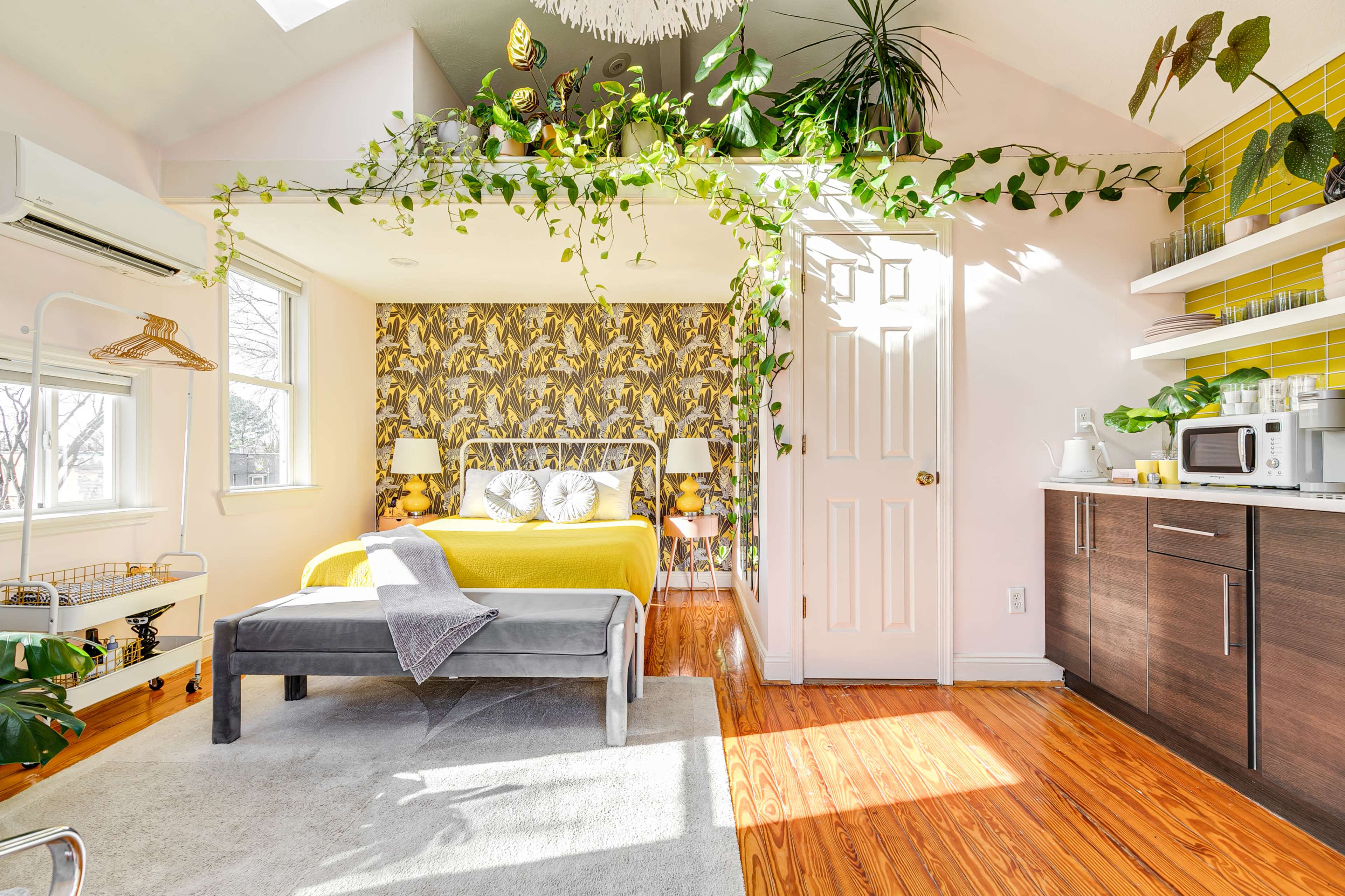 The image shows a bright bedroom with a yellow bedspread, large windows, and indoor plants cascading from the ceiling.