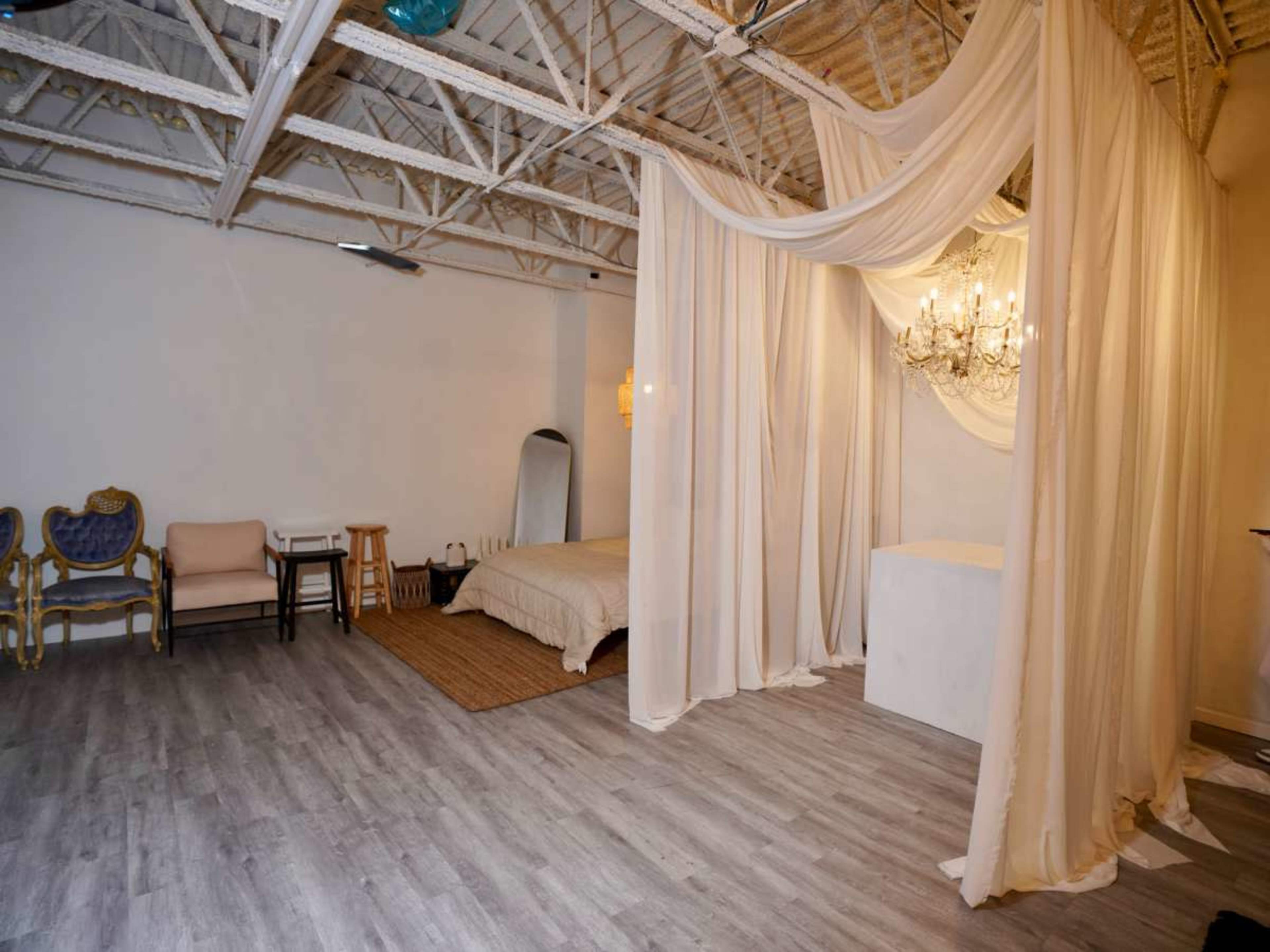 The image shows a spacious room with wooden flooring, a bed draped in neutral fabric, a chandelier, and a few seated areas.
