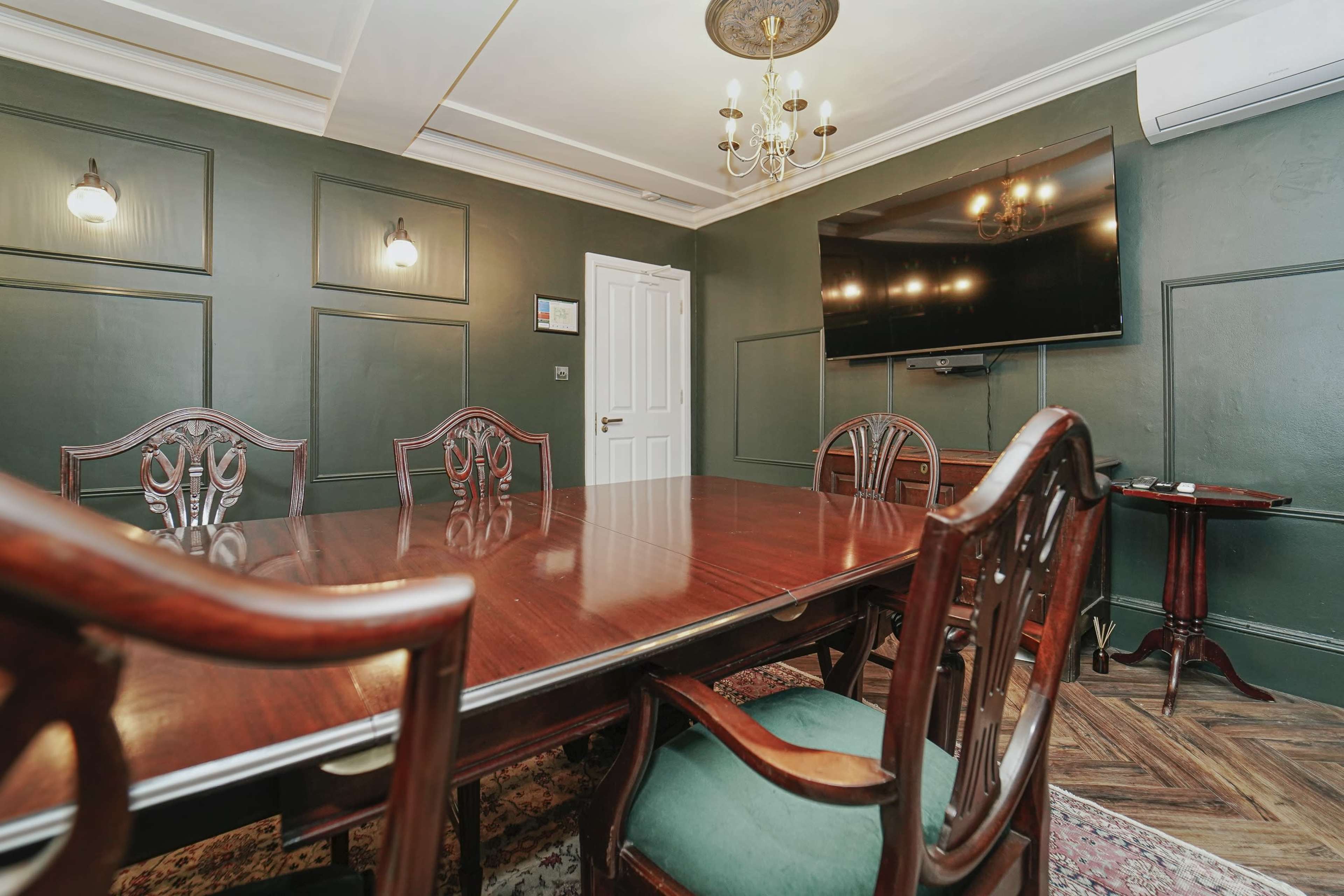 A wooden conference table is set in a room with dark green walls, featuring a television mounted on one wall and antique-style chairs around the table.