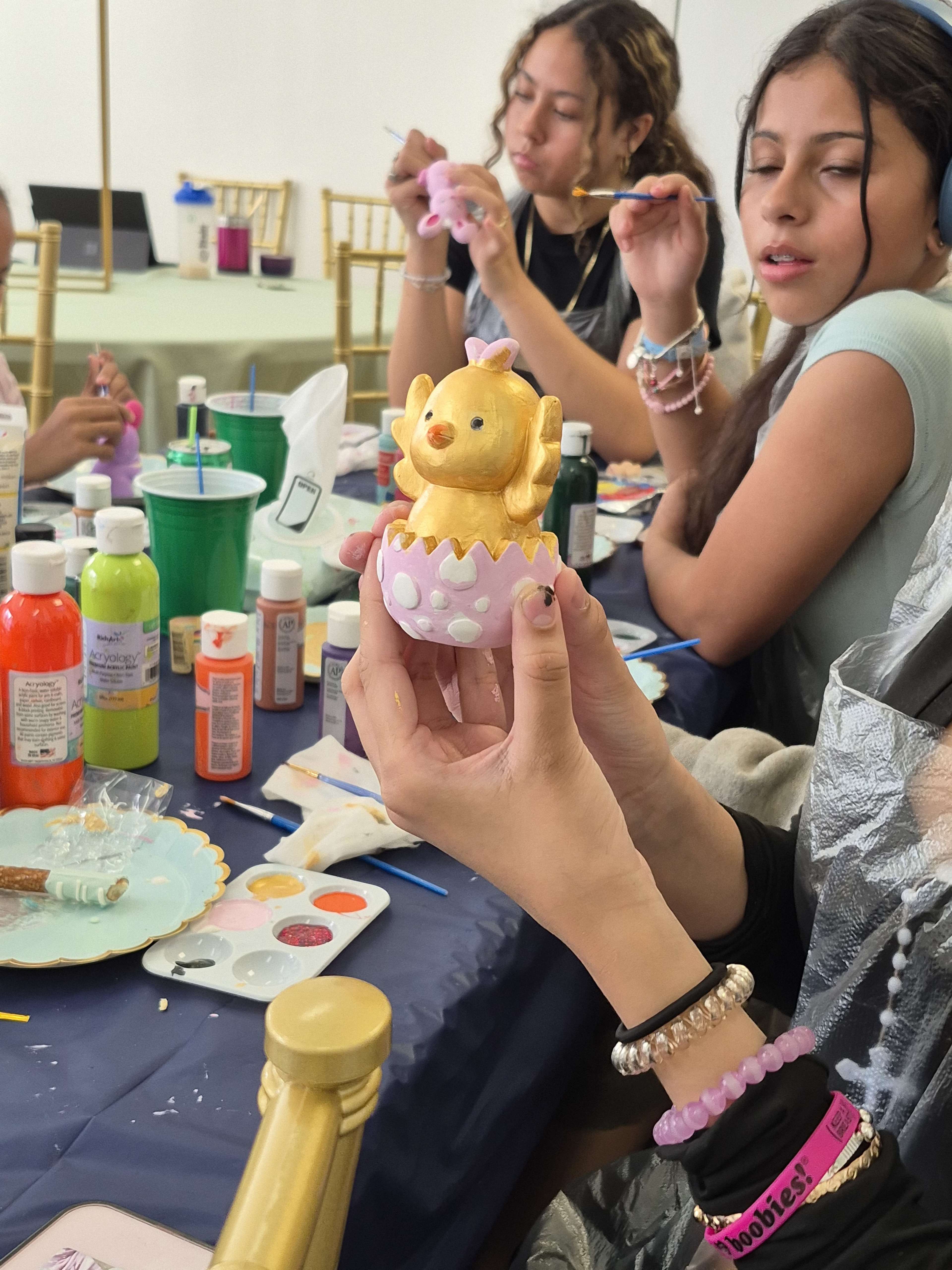 A person holds a yellow painted duck figurine emerging from a pink egg while others around them are engaged in painting at a craft table.