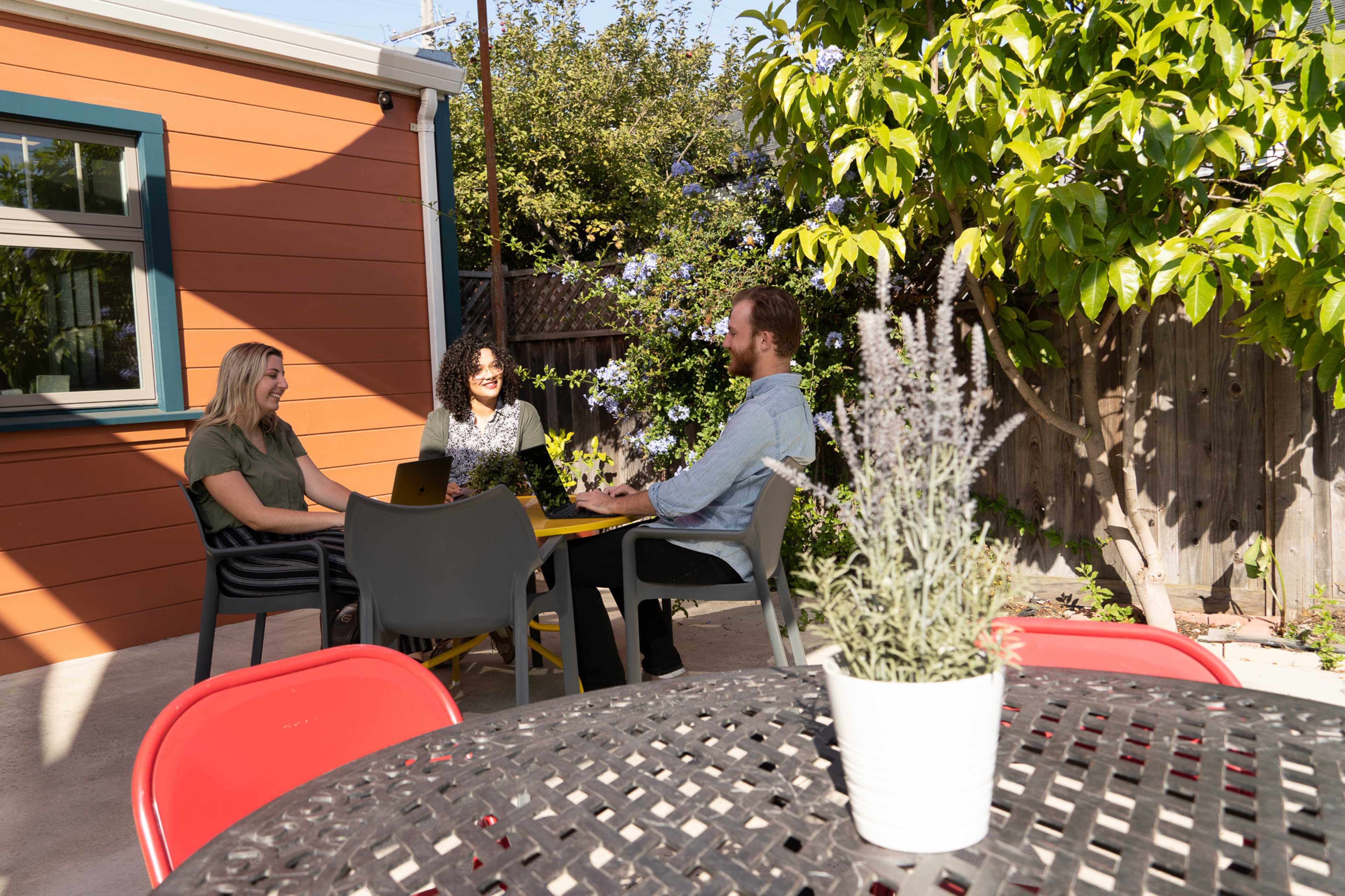 Three people sit at a yellow table in a backyard, discussing while surrounded by plants and a colorful wall.