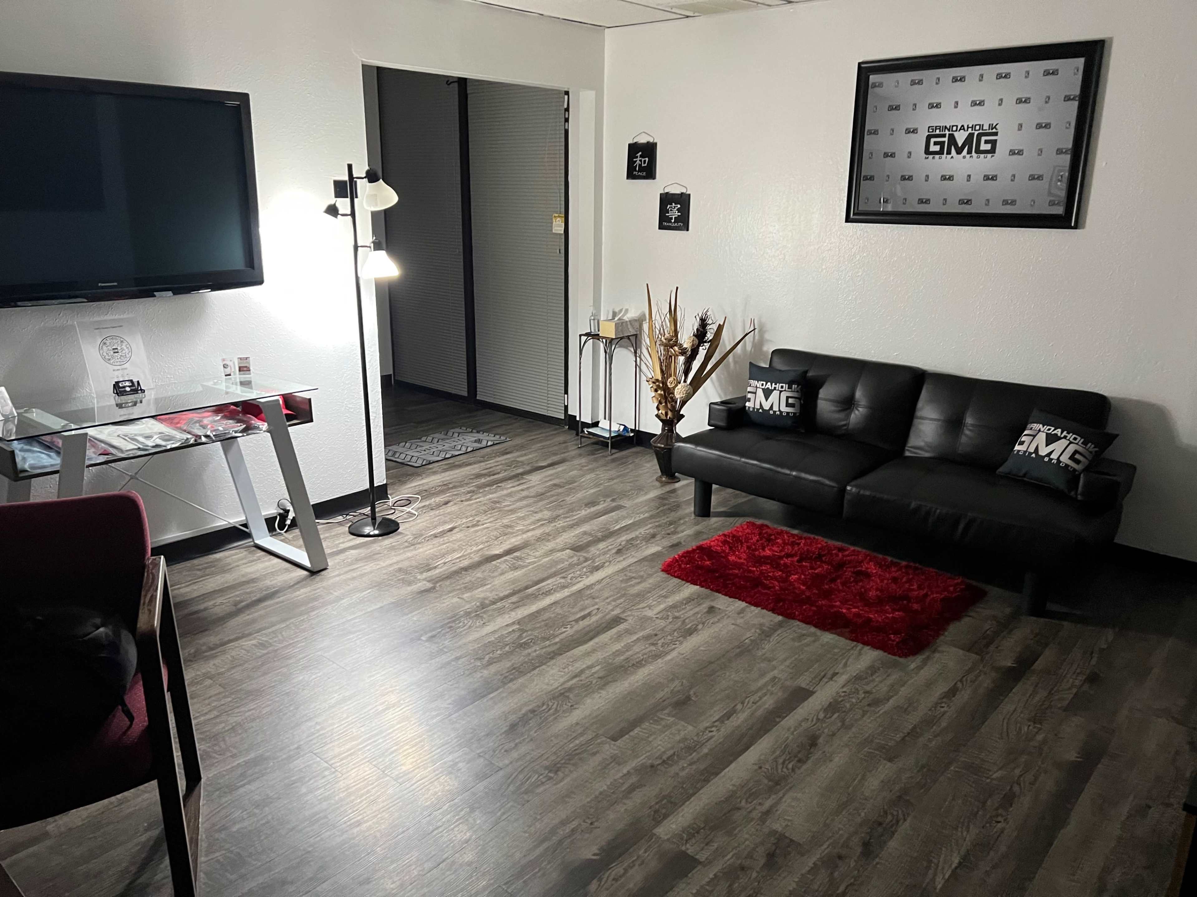 A modern office space with a black couch, a television, and a lamp, featuring a wooden floor and a red rug.