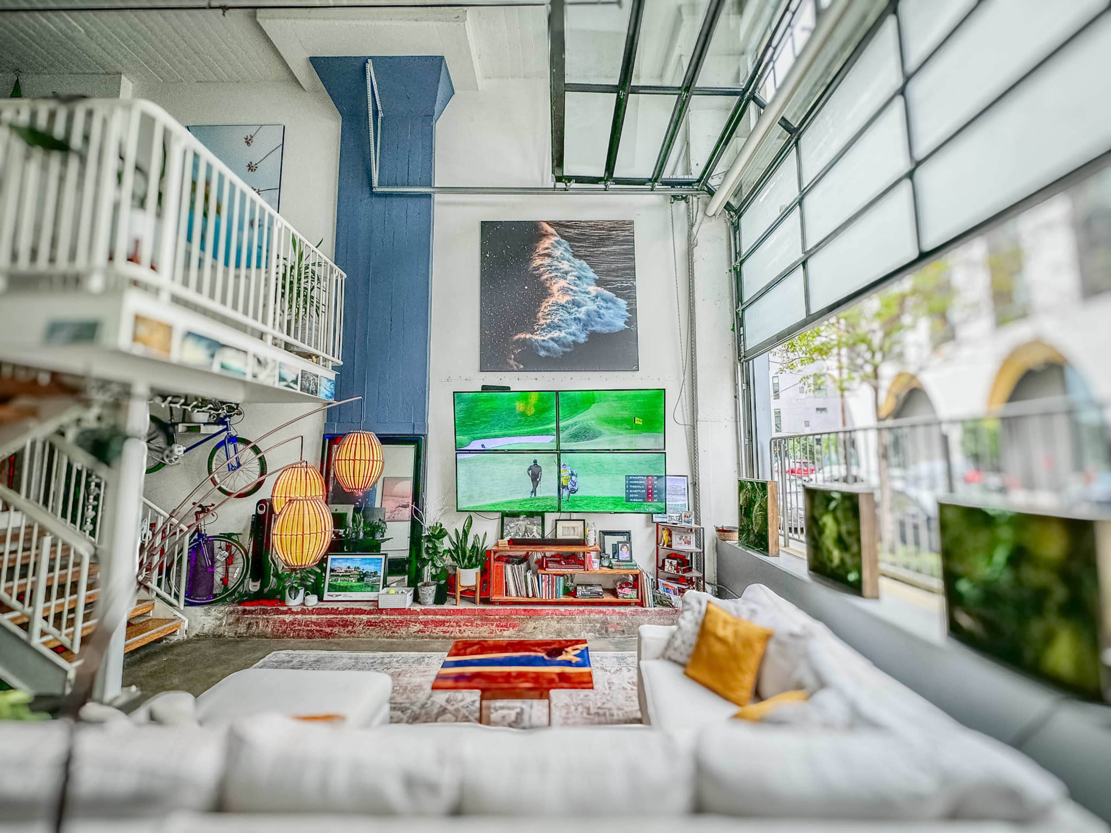 The image shows a large, modern living space with an open layout featuring a white sofa, a colorful rug, and two large screens displaying a golf game, complemented by art on the walls and bicycles in the background.