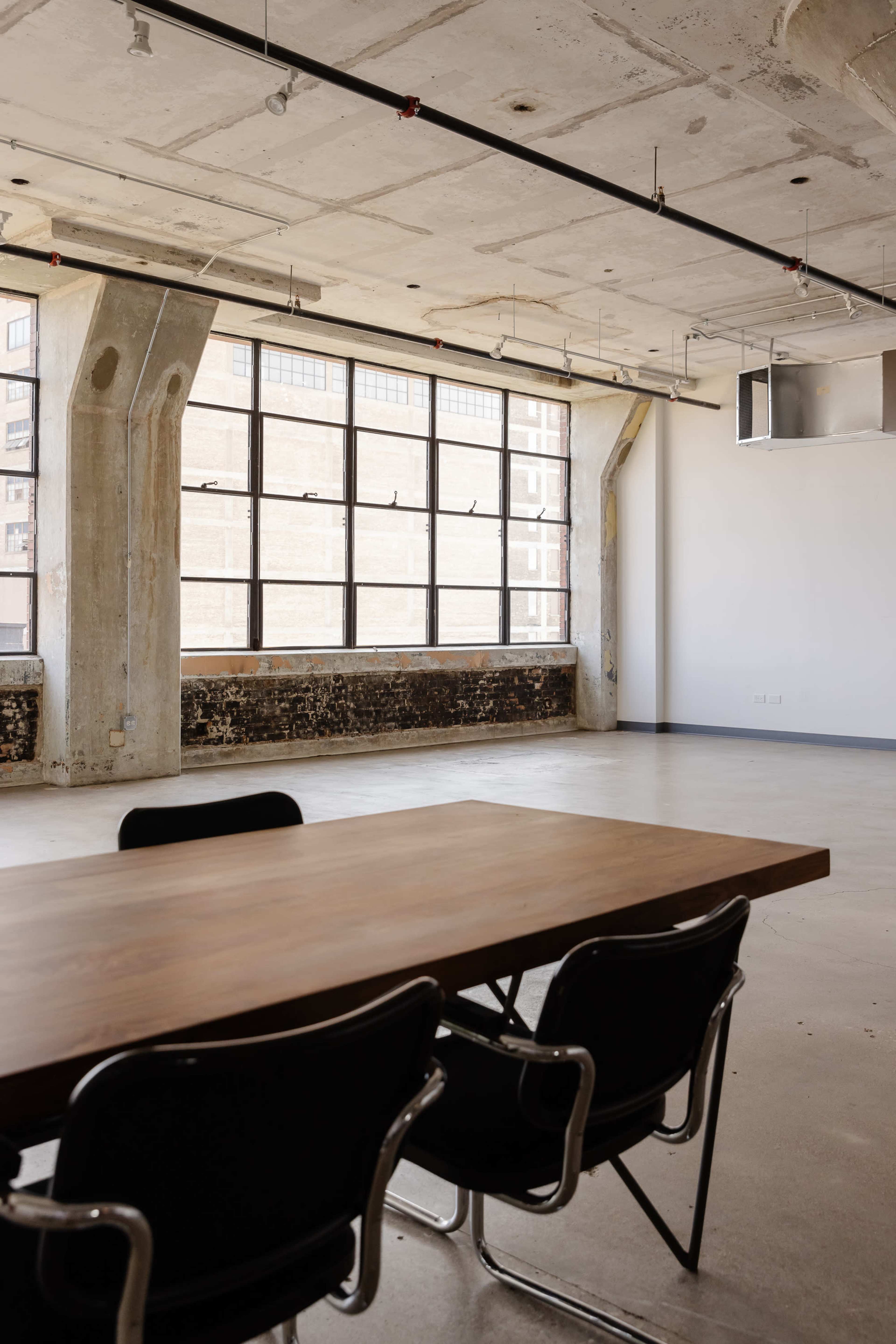 An empty meeting room features a wooden table surrounded by four black chairs, with large industrial windows letting in natural light.