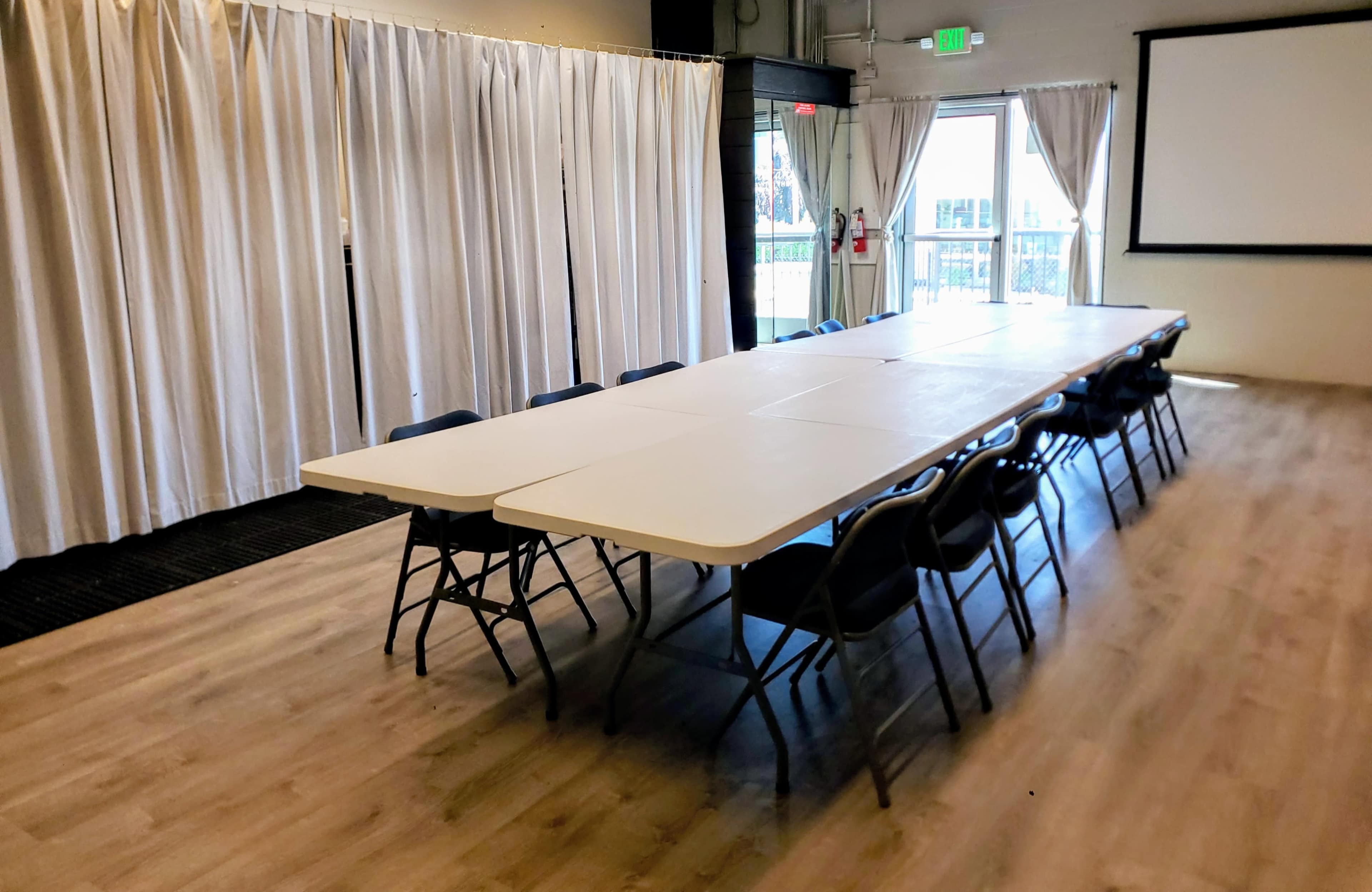 A long table surrounded by chairs is set up in a room with white curtains and a large screen at one end.
