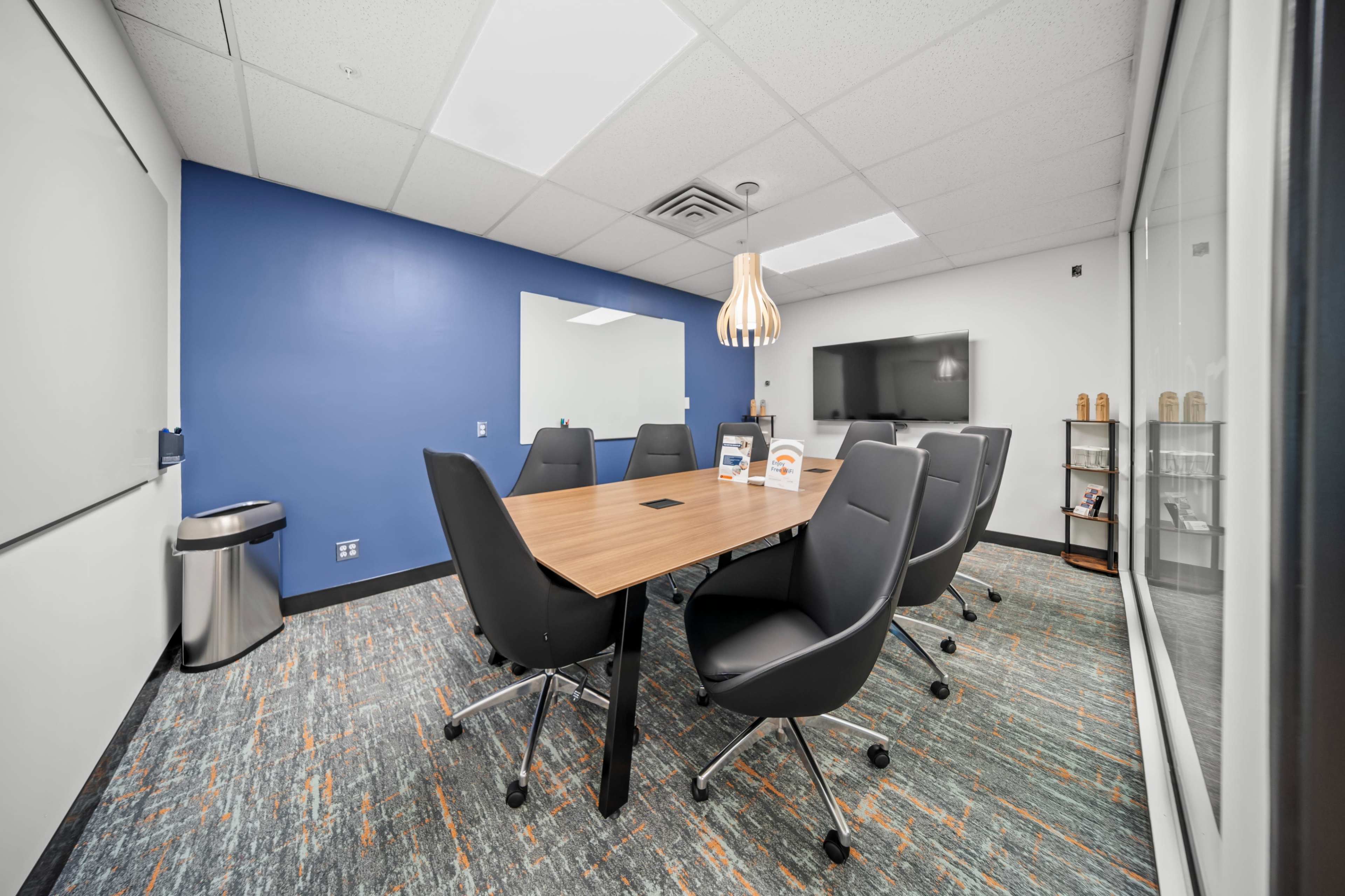 The image shows a modern conference room with a wooden table, black chairs, a large screen, and a blue accent wall.