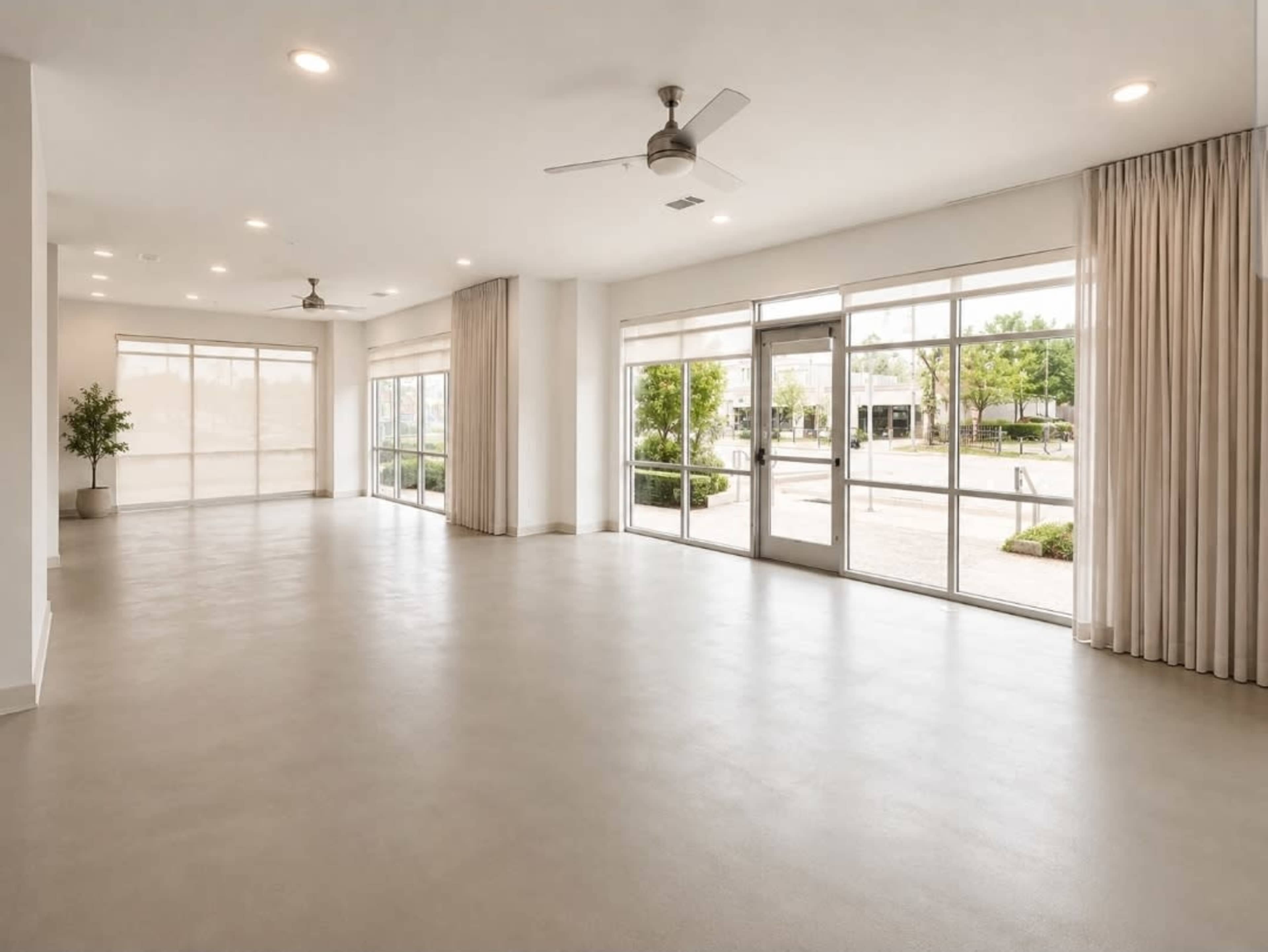 A spacious, well-lit room features large windows and minimalist decor, overlooking an outdoor area with greenery.