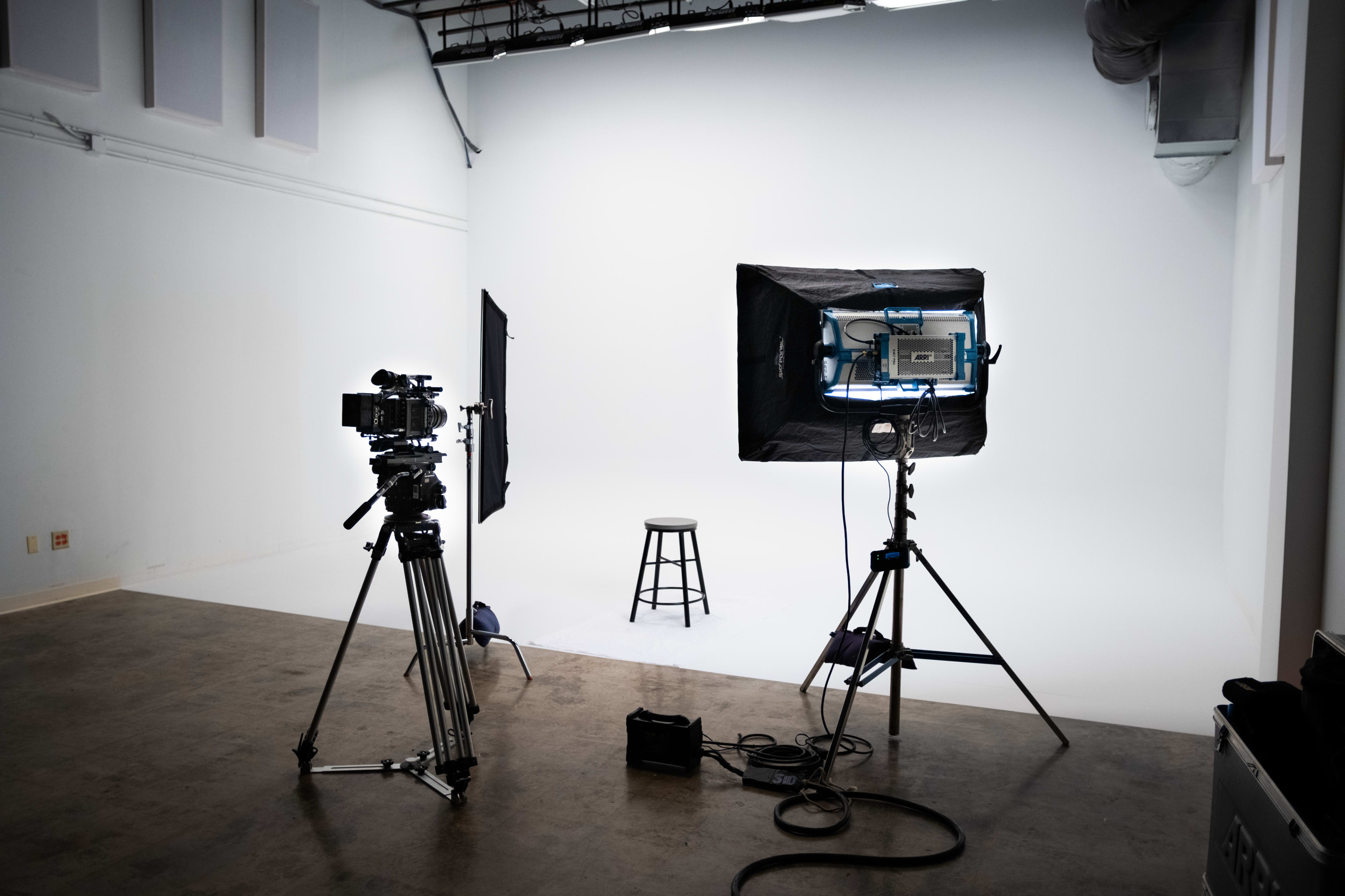A film studio setup with two cameras and lighting equipment facing an empty stool against a white backdrop.