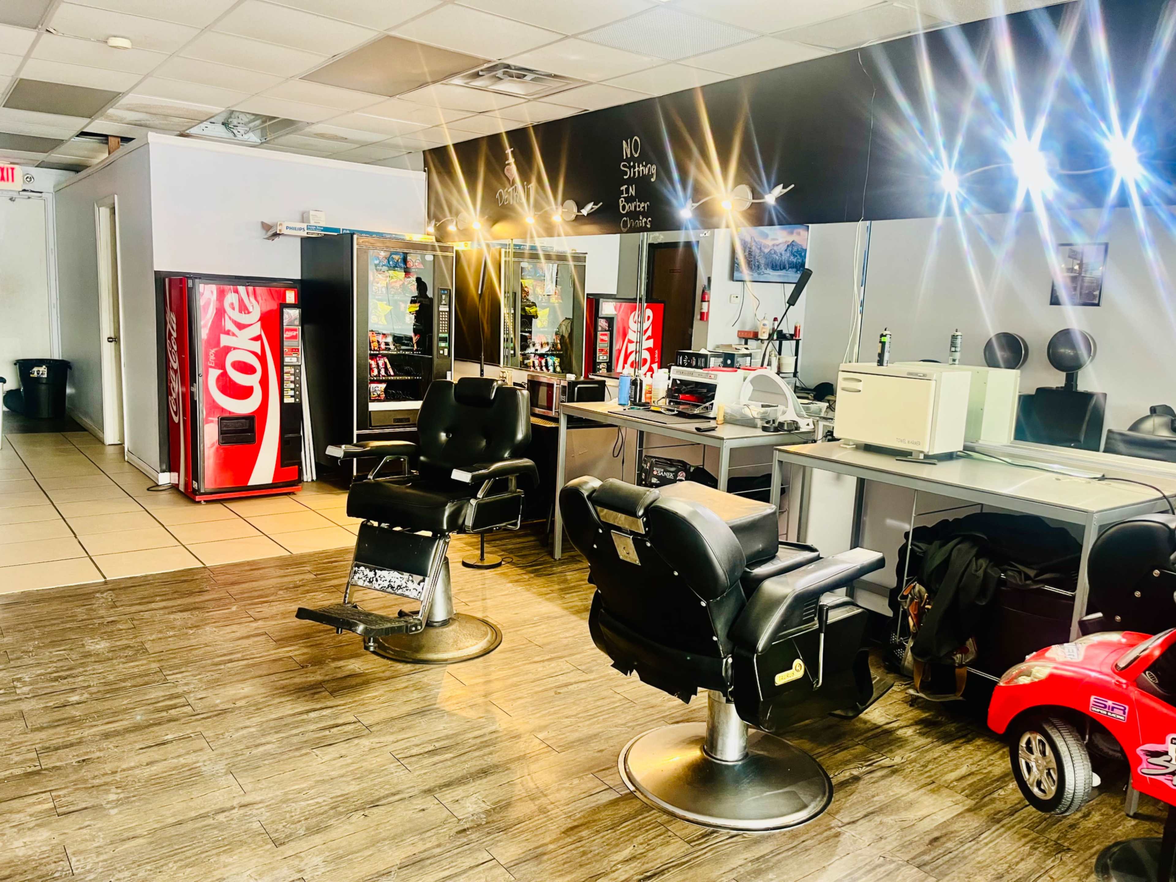 The image shows a hair salon interior with two black barber chairs, illuminated mirrors, and a Coca-Cola vending machine in the background.