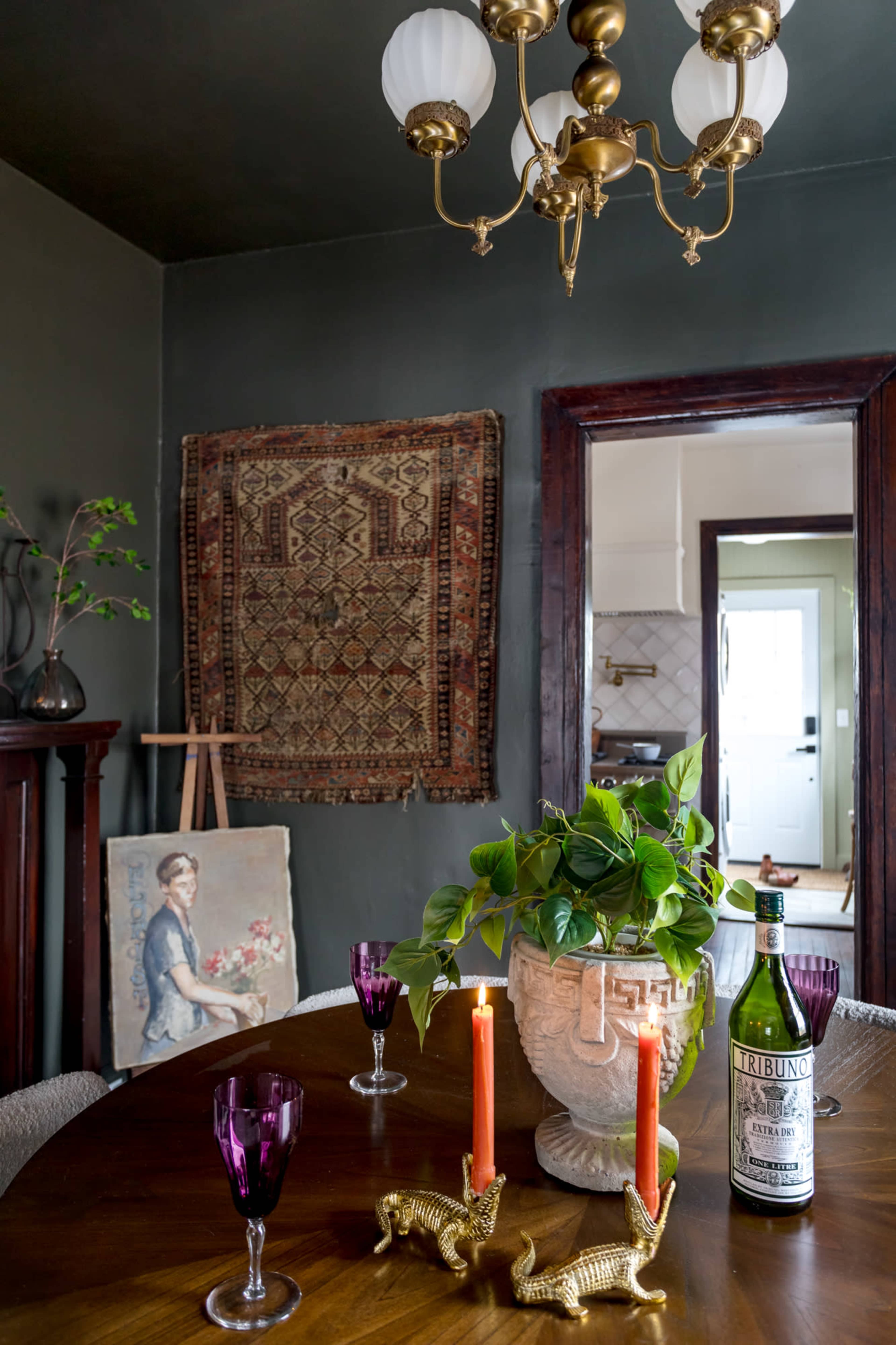 A dining table is set with two purple glasses, two candles, and a decorative plant, surrounded by patterned fabric and artwork against olive-green walls.