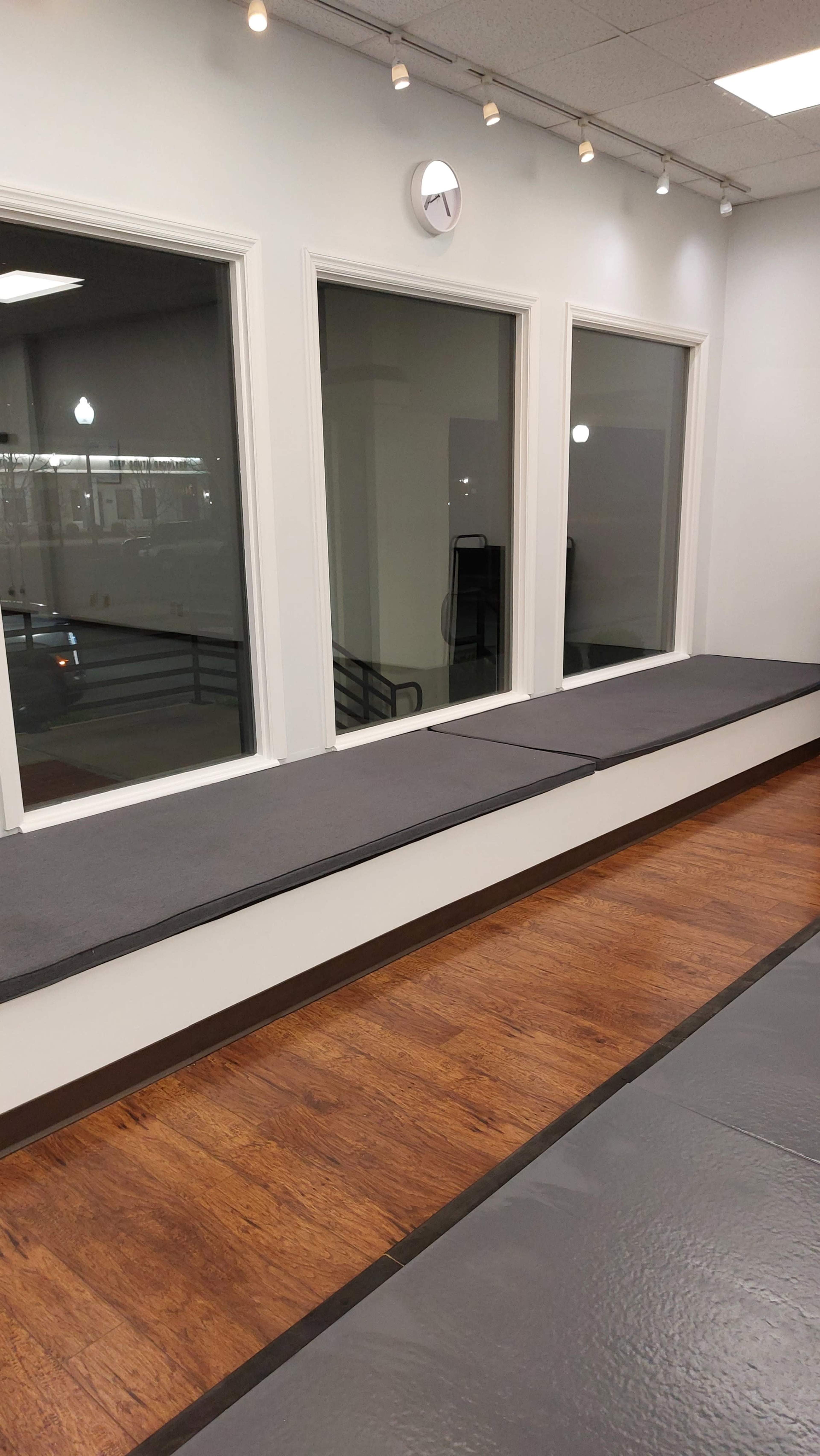 The image shows an interior space with large windows, a long bench along the wall, and a wooden floor covered by gray mats.