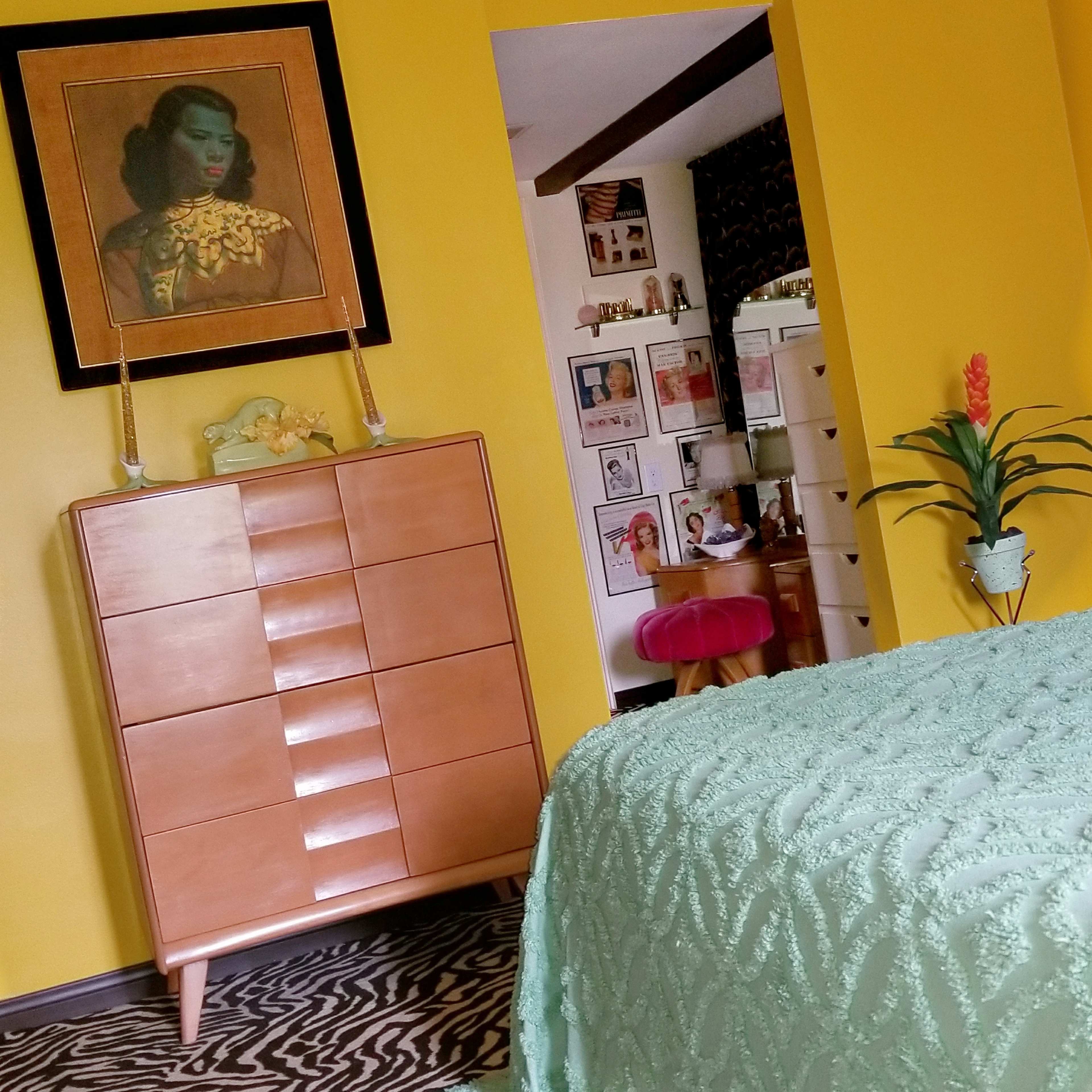 The image shows a brightly painted yellow room featuring a mid-century dresser, a vibrant green bedspread, and an adjoining room with framed artwork and a desk.