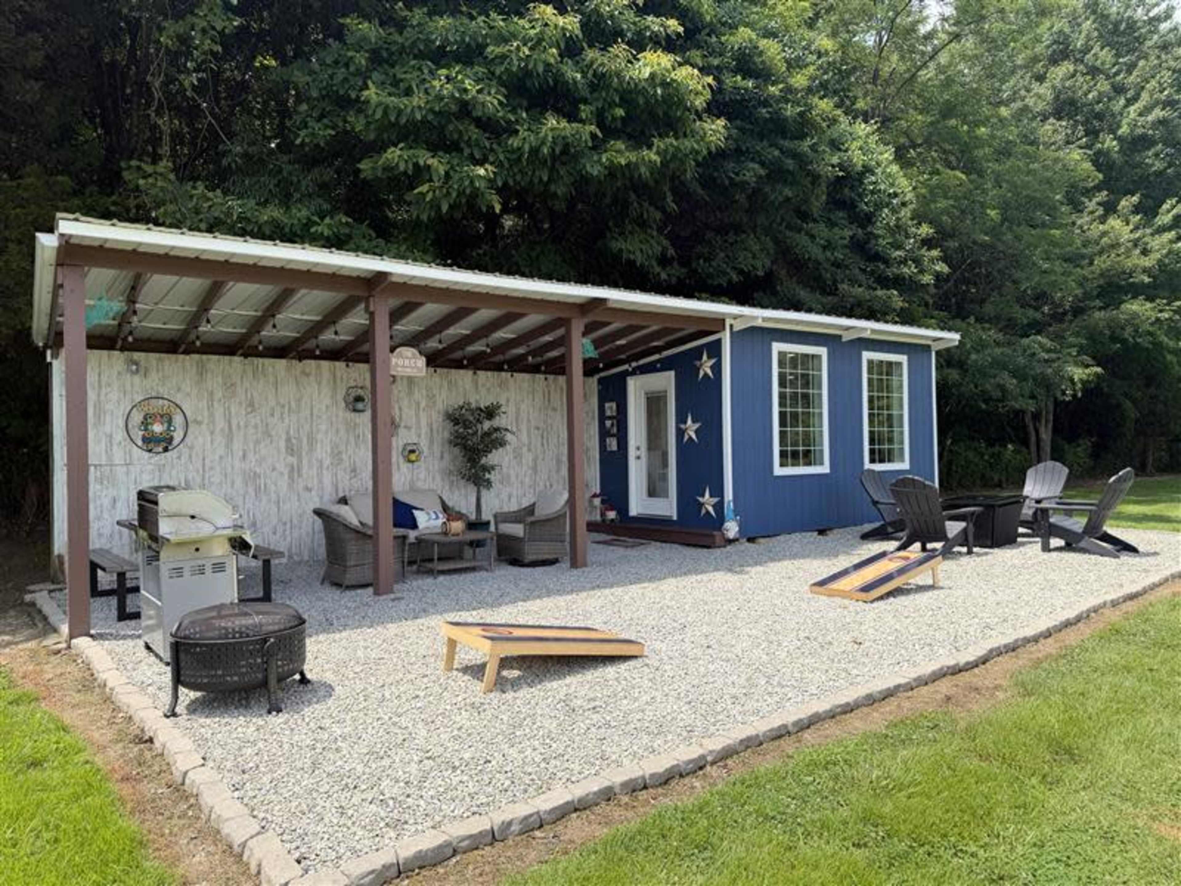 The image shows a blue building with a covered porch area featuring seating and a grill, surrounded by a gravel yard and lawn.