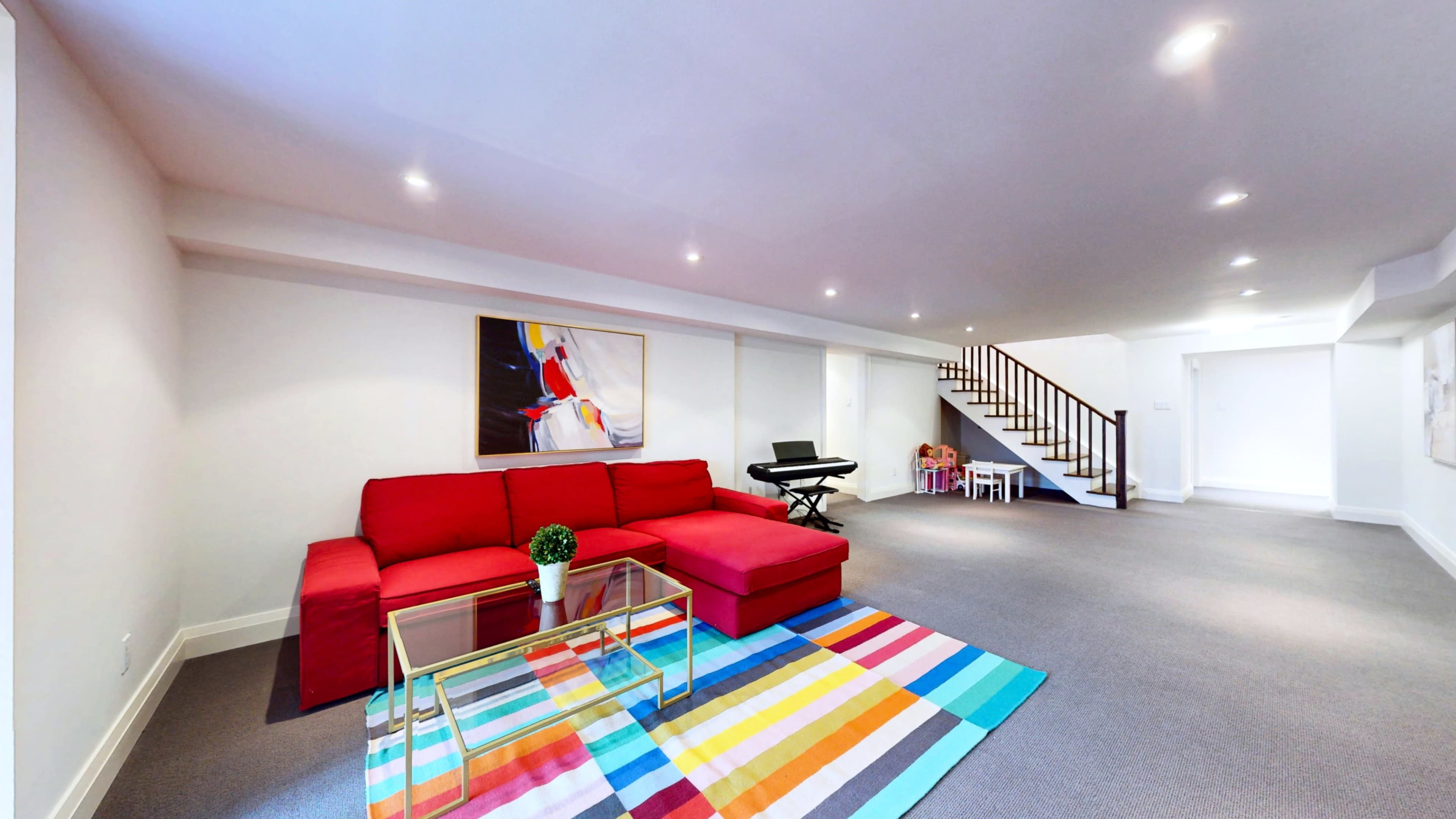 The image shows a modern basement living area with a red sofa, a colorful striped rug, and a staircase in the background.