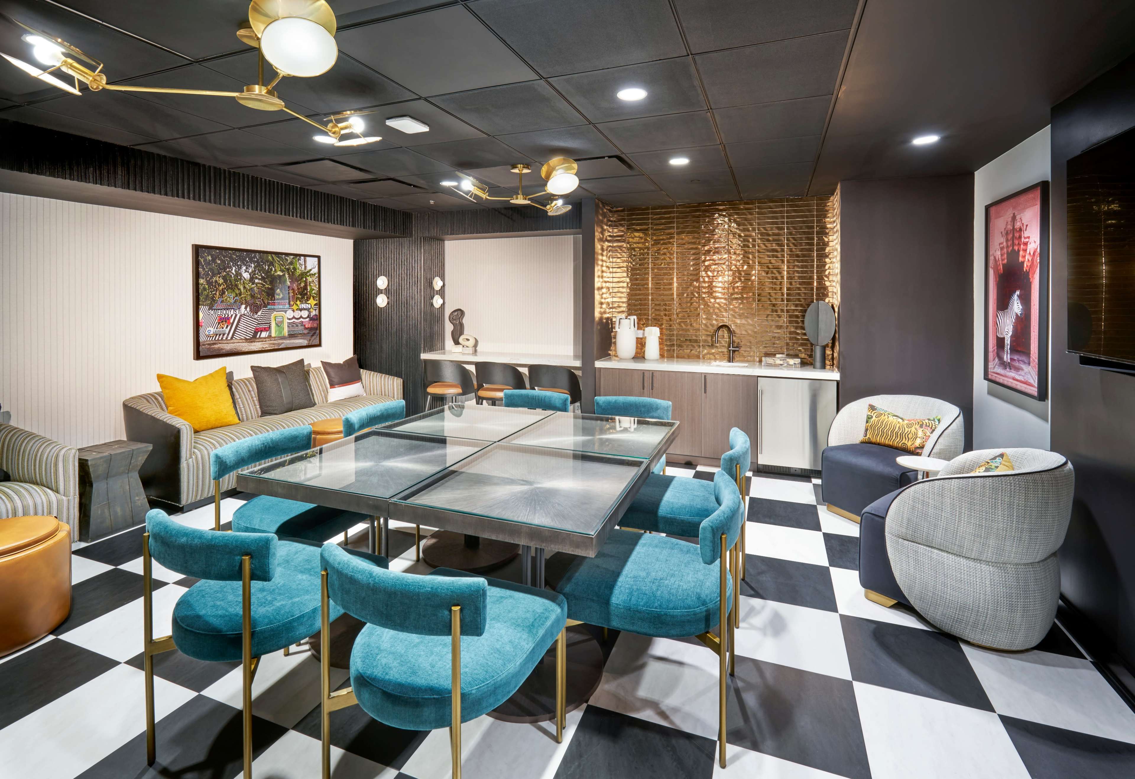 The image depicts a modern lounge area featuring a glass-top table surrounded by blue chairs, a kitchenette with a gold backsplash, and various decorative elements.
