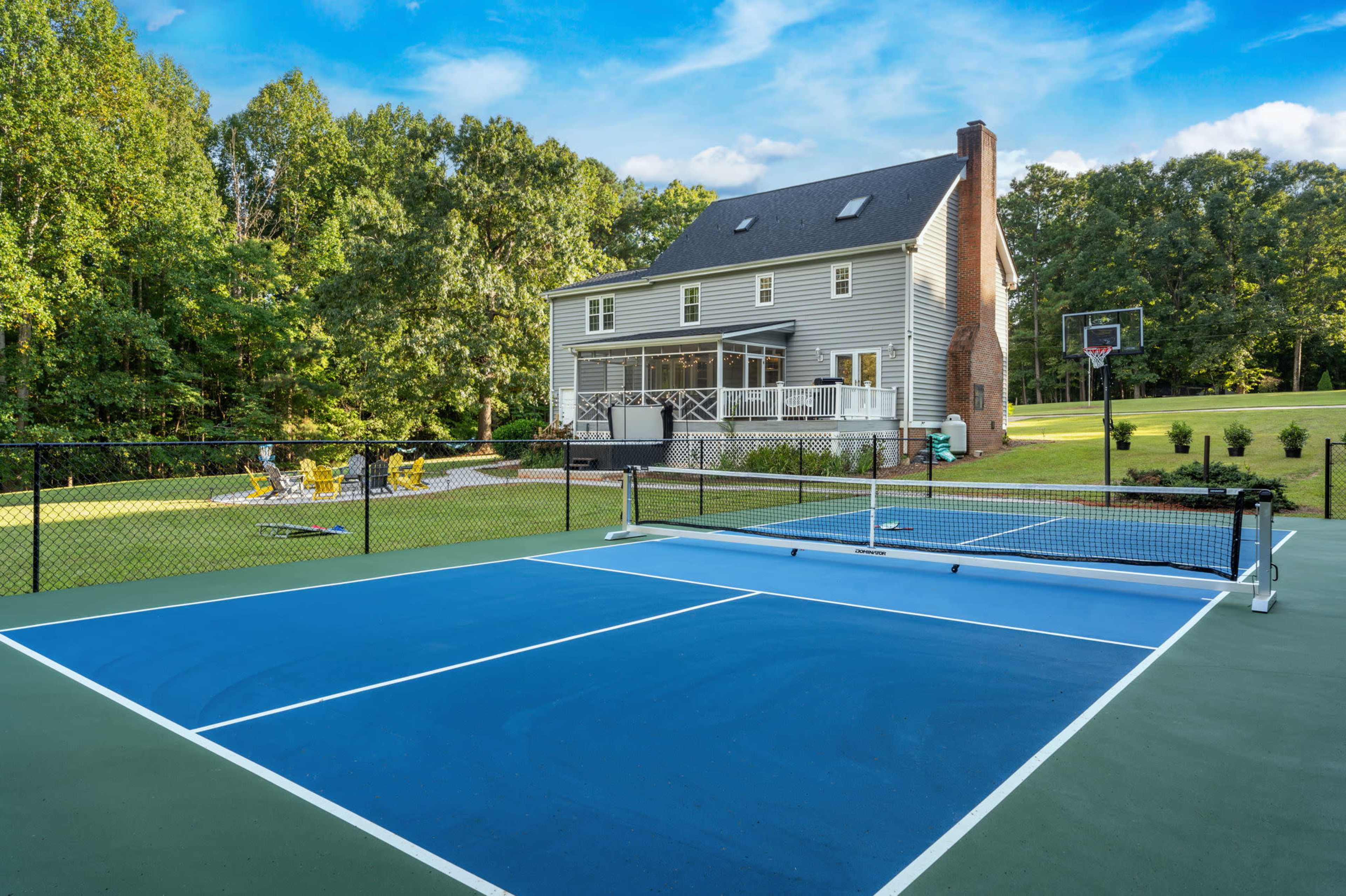 The image shows a well-maintained home with a large green lawn and a fenced basketball court, alongside a blue tennis court.
