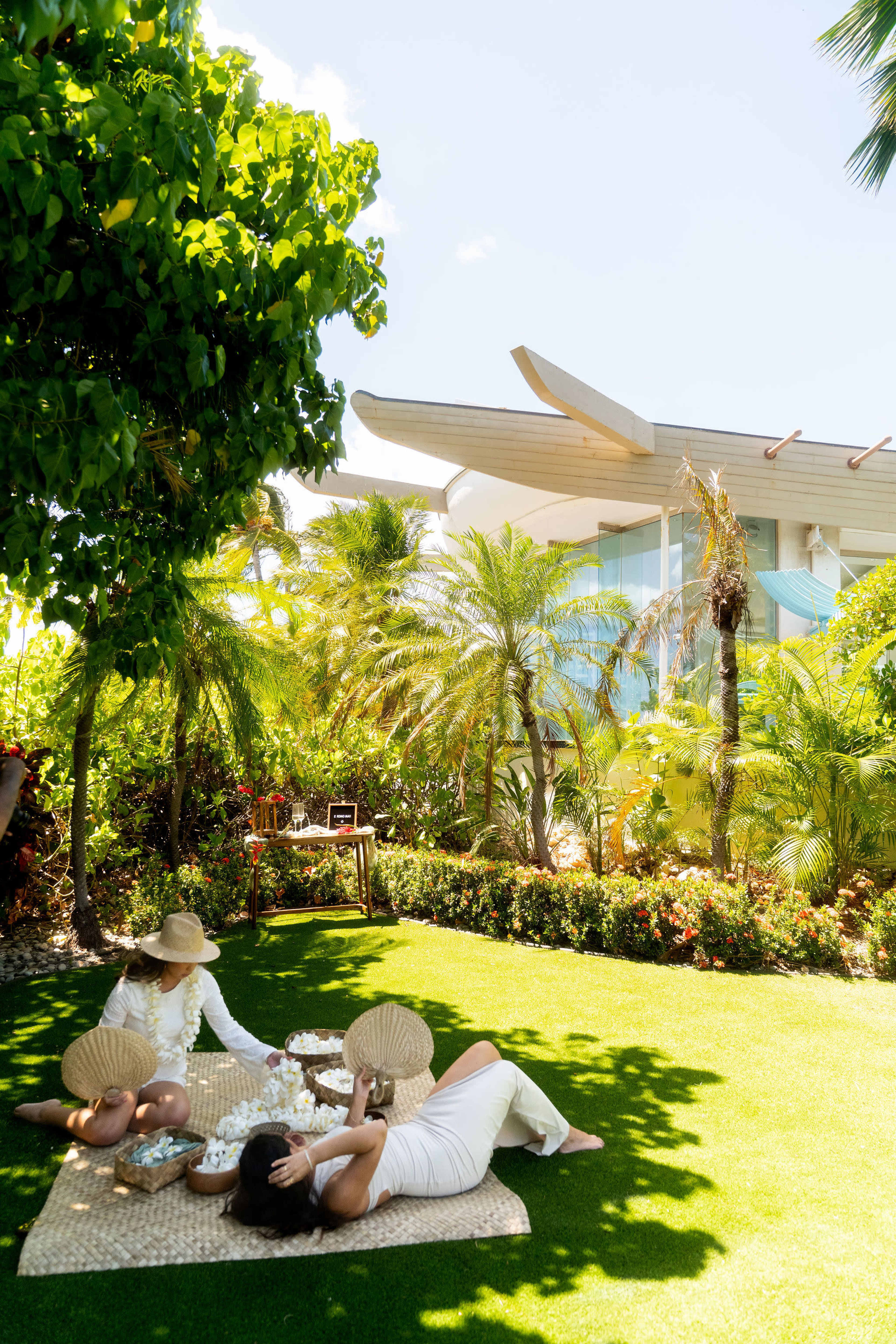 Two women relax on a blanket in a lush garden, surrounded by tropical plants and a modern house in the background.