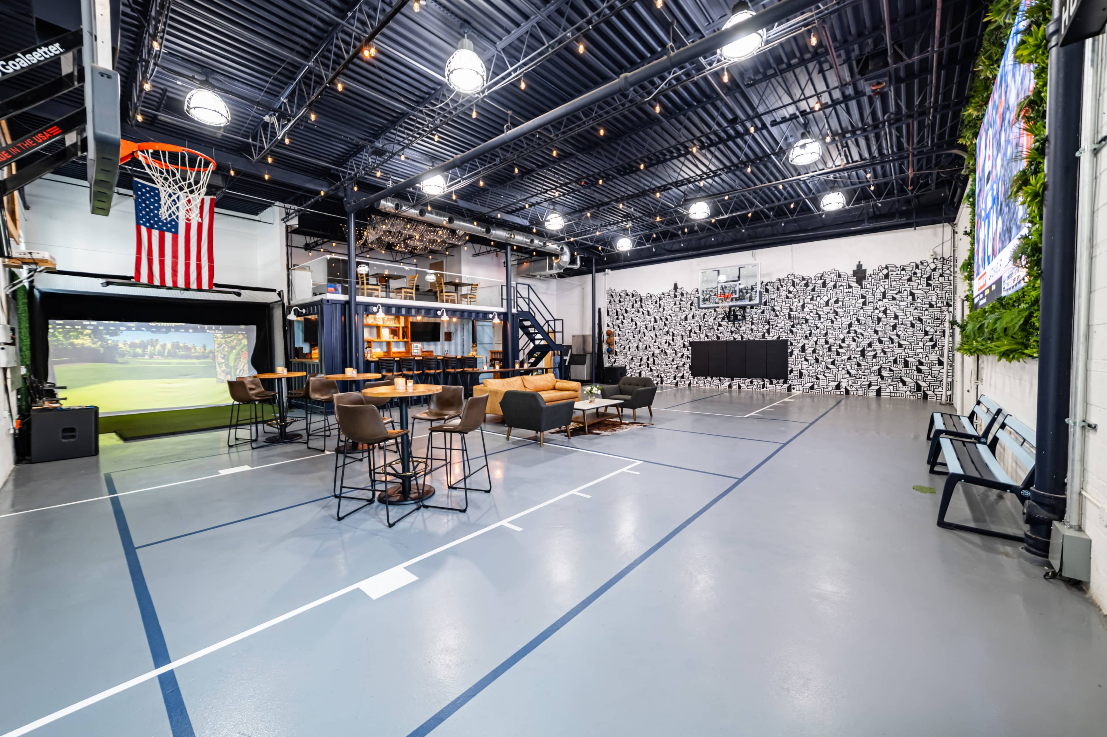 A spacious entertainment area featuring a basketball hoop, a golf simulator screen, a large TV, seating areas, and modern decor with an American flag hanging from the ceiling.