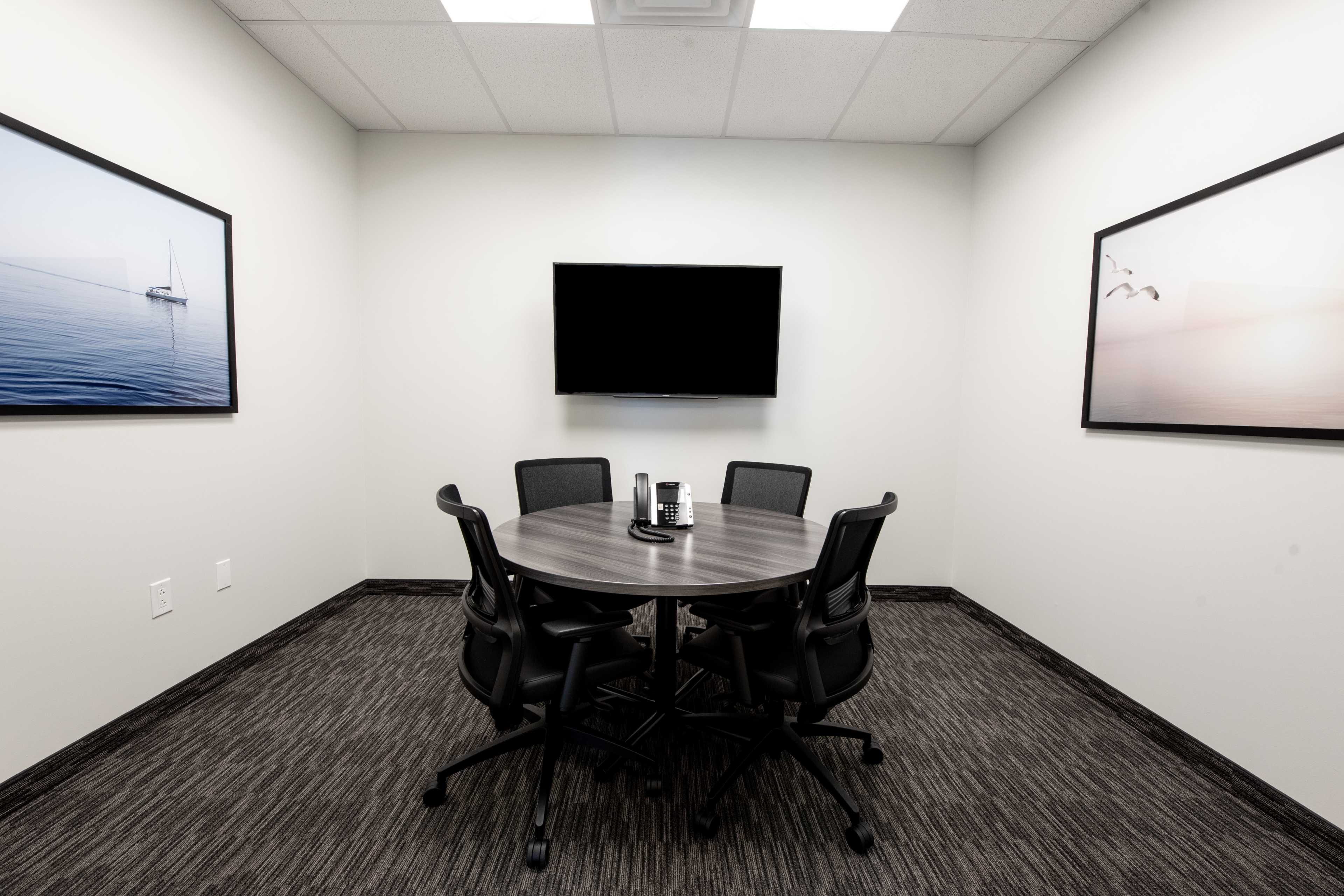 A small conference room features a round table with four chairs, a wall-mounted television, and two framed photos on the walls.