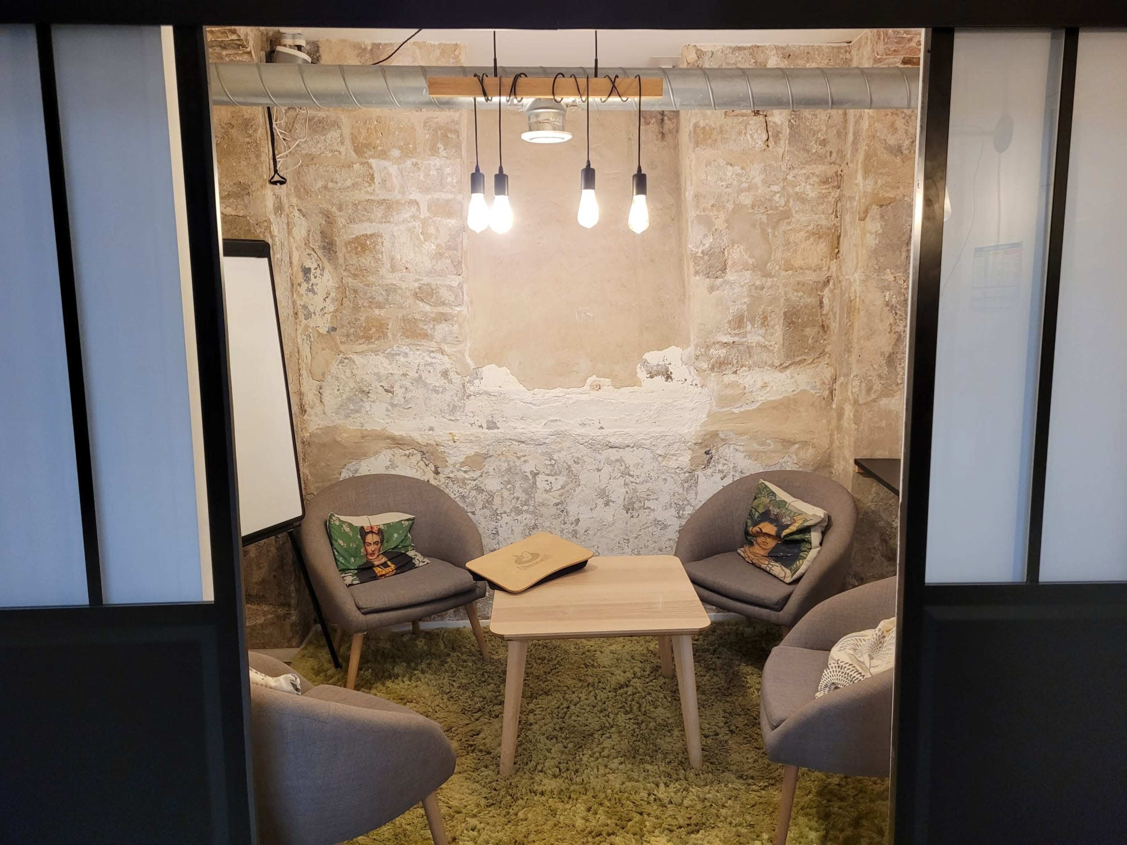 A small meeting room with four gray chairs arranged around a wooden table, illuminated by hanging light bulbs against a stone wall.