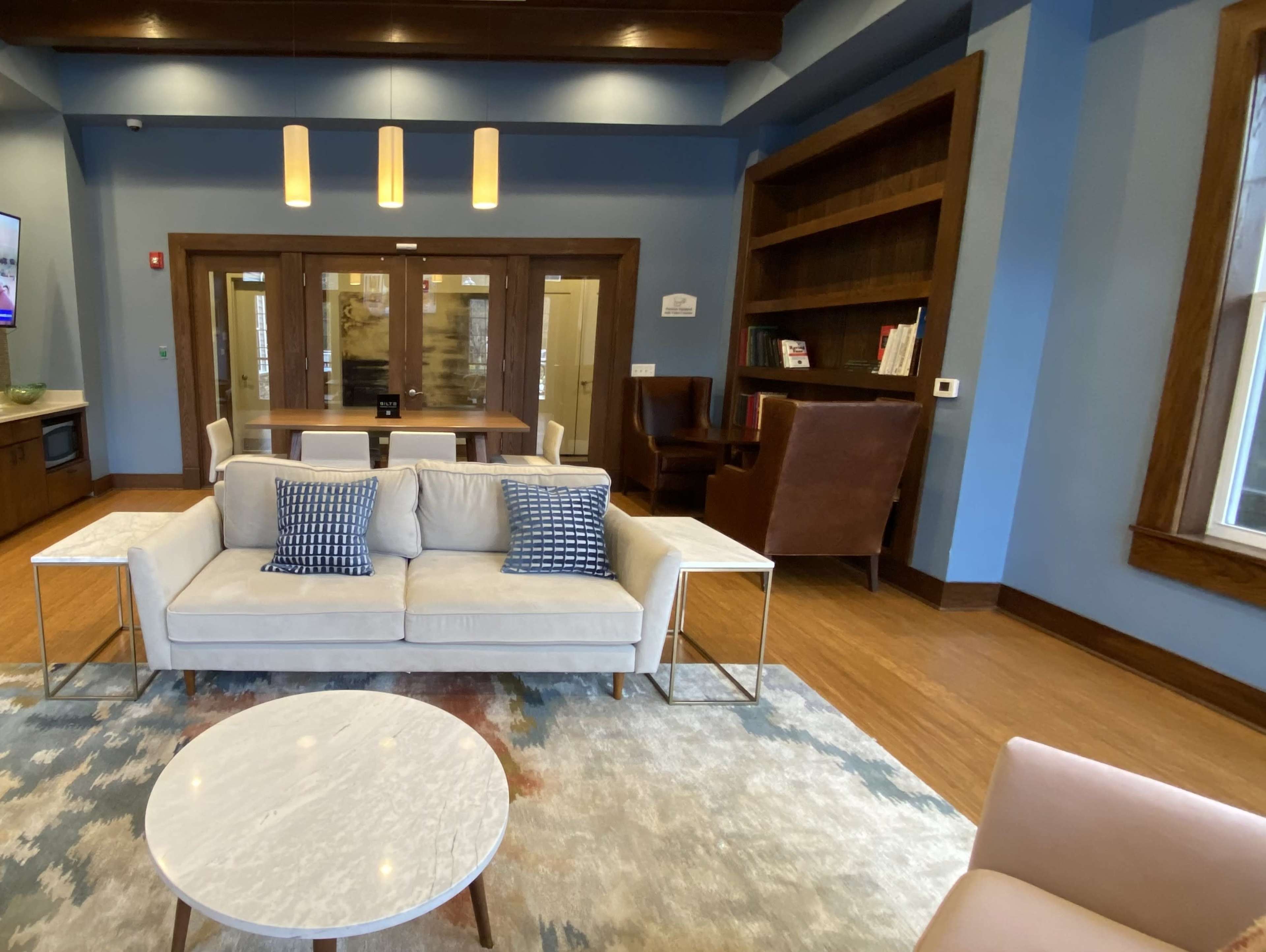A modern seating area features a light sofa with decorative pillows, a marble-top coffee table, and wooden bookshelves against blue walls.