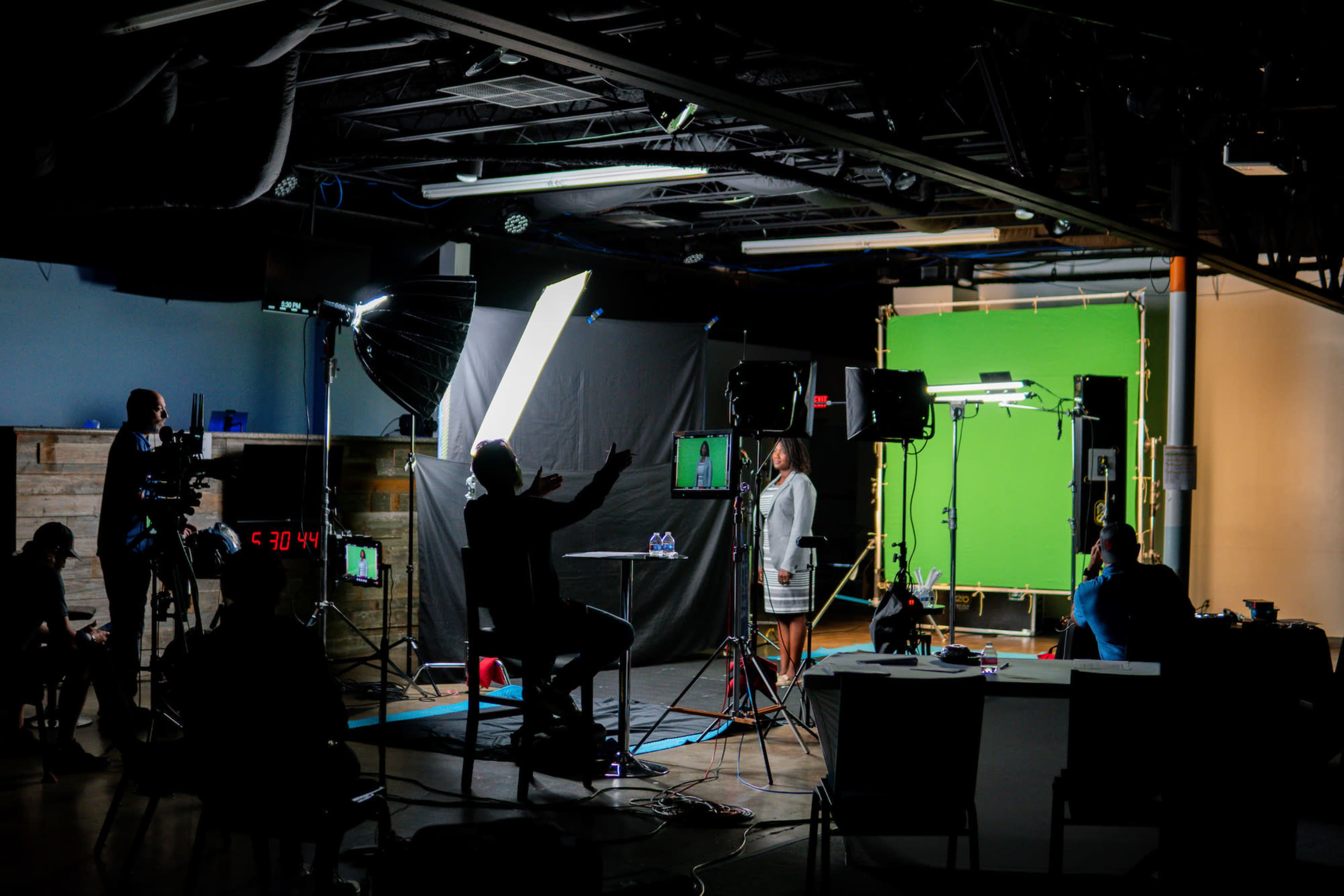 A video production setup features a woman in front of a green screen, flanked by crew members using cameras and lighting equipment in a dimly lit studio.