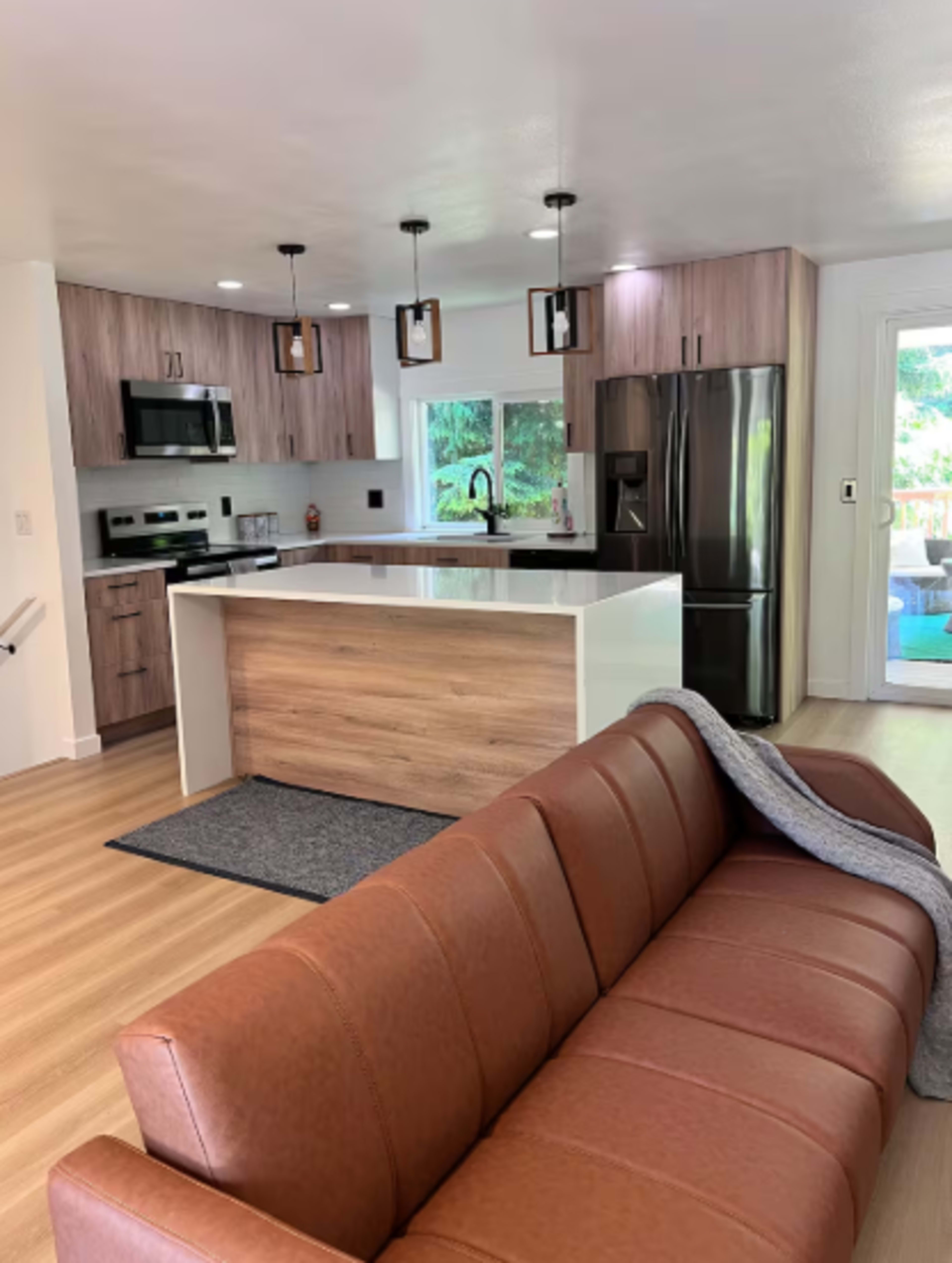 The image shows a modern kitchen with wooden cabinetry, stainless steel appliances, and a large island, adjacent to a living area featuring a brown leather sofa.
