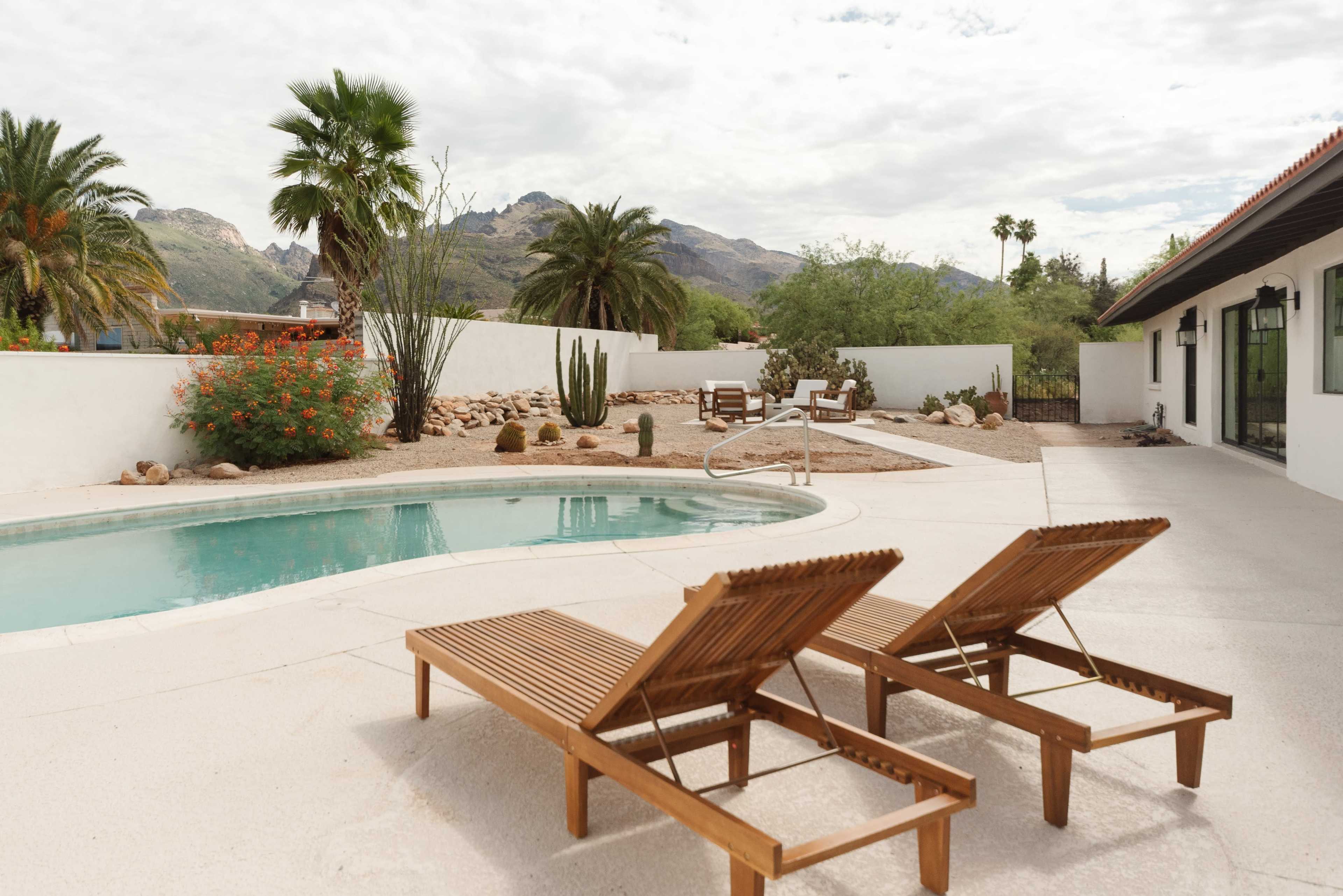 A backyard with a swimming pool, two wooden lounge chairs, and desert landscaping, framed by mountains and palm trees in the background.