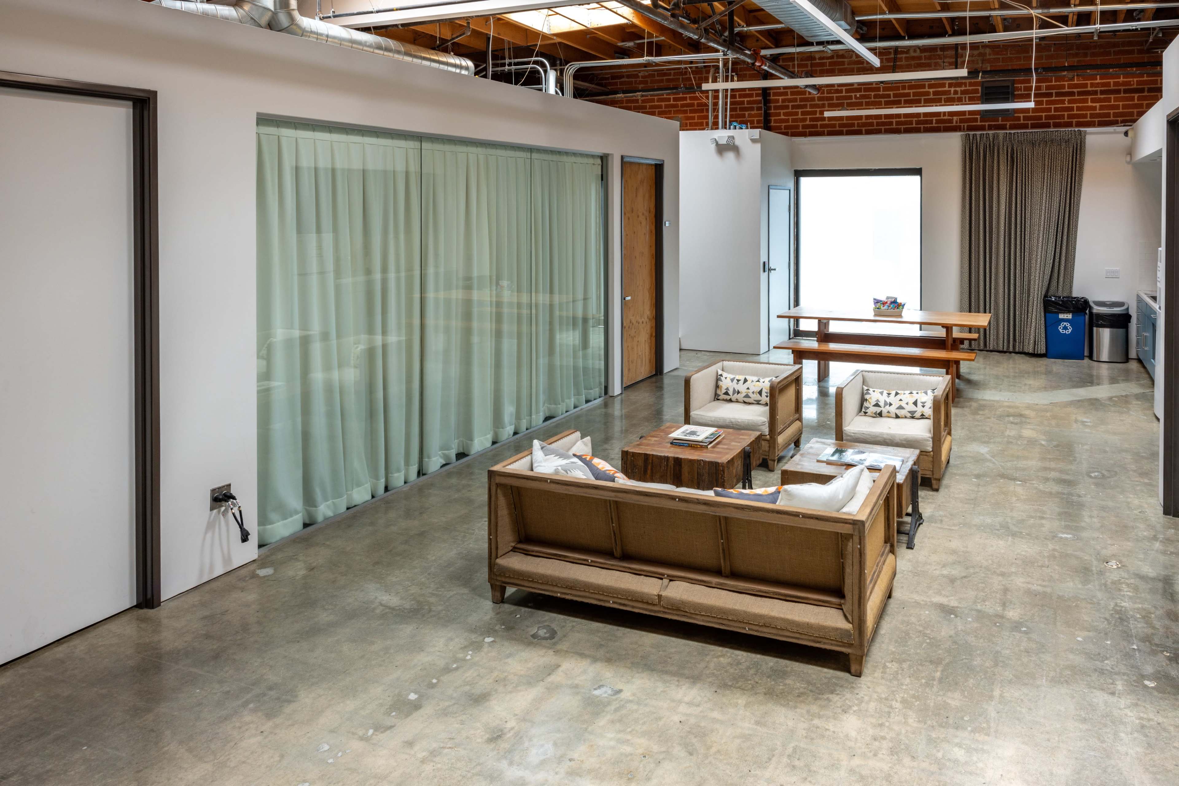 The image shows a spacious room with a concrete floor, featuring a seating area with four chairs and a low table, alongside a large window covered with sheer curtains.