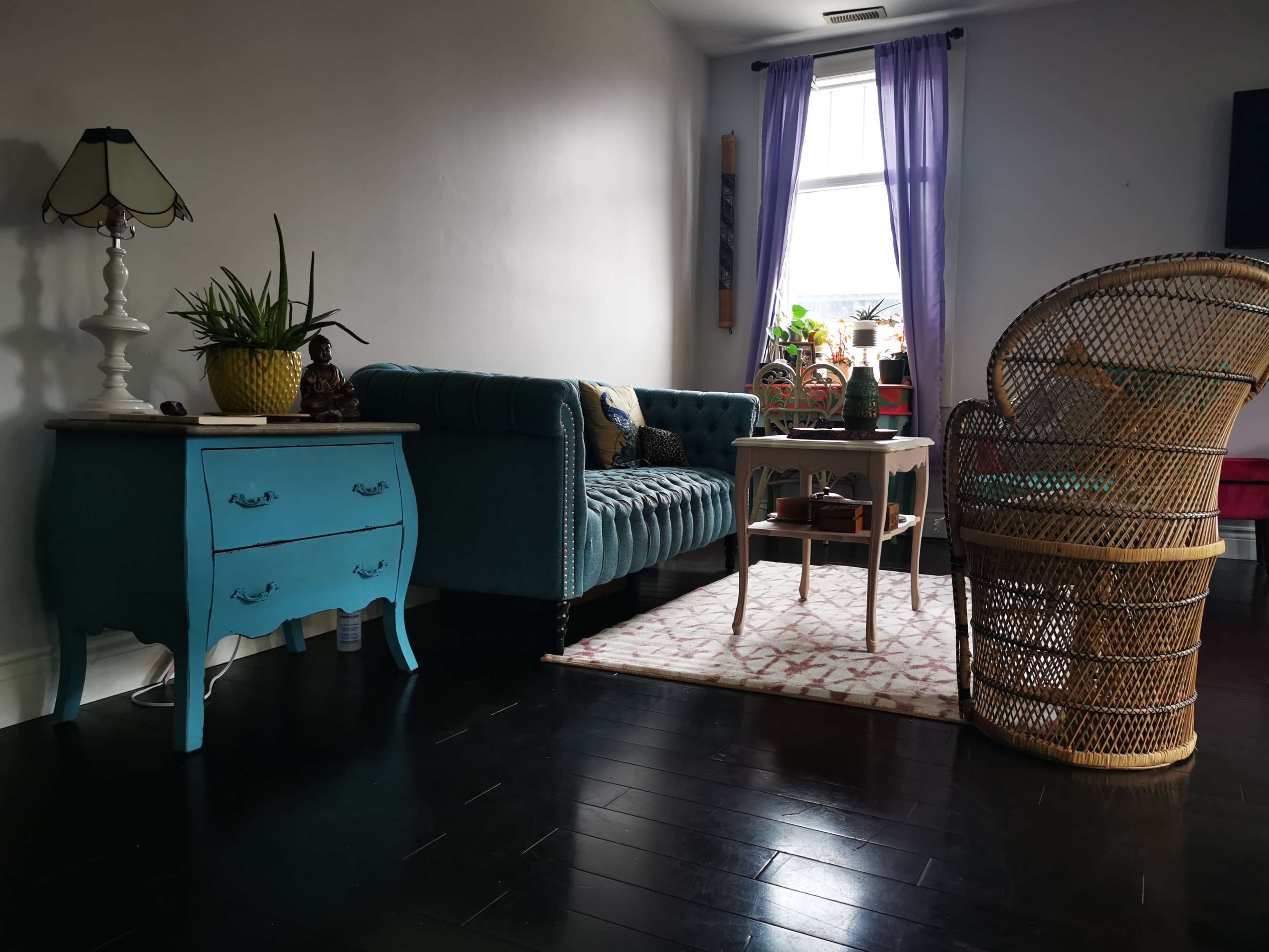 The image shows a living room featuring a teal velvet couch, a small coffee table, a wicker chair, and a blue-painted chest of drawers, all illuminated by natural light from a nearby window.