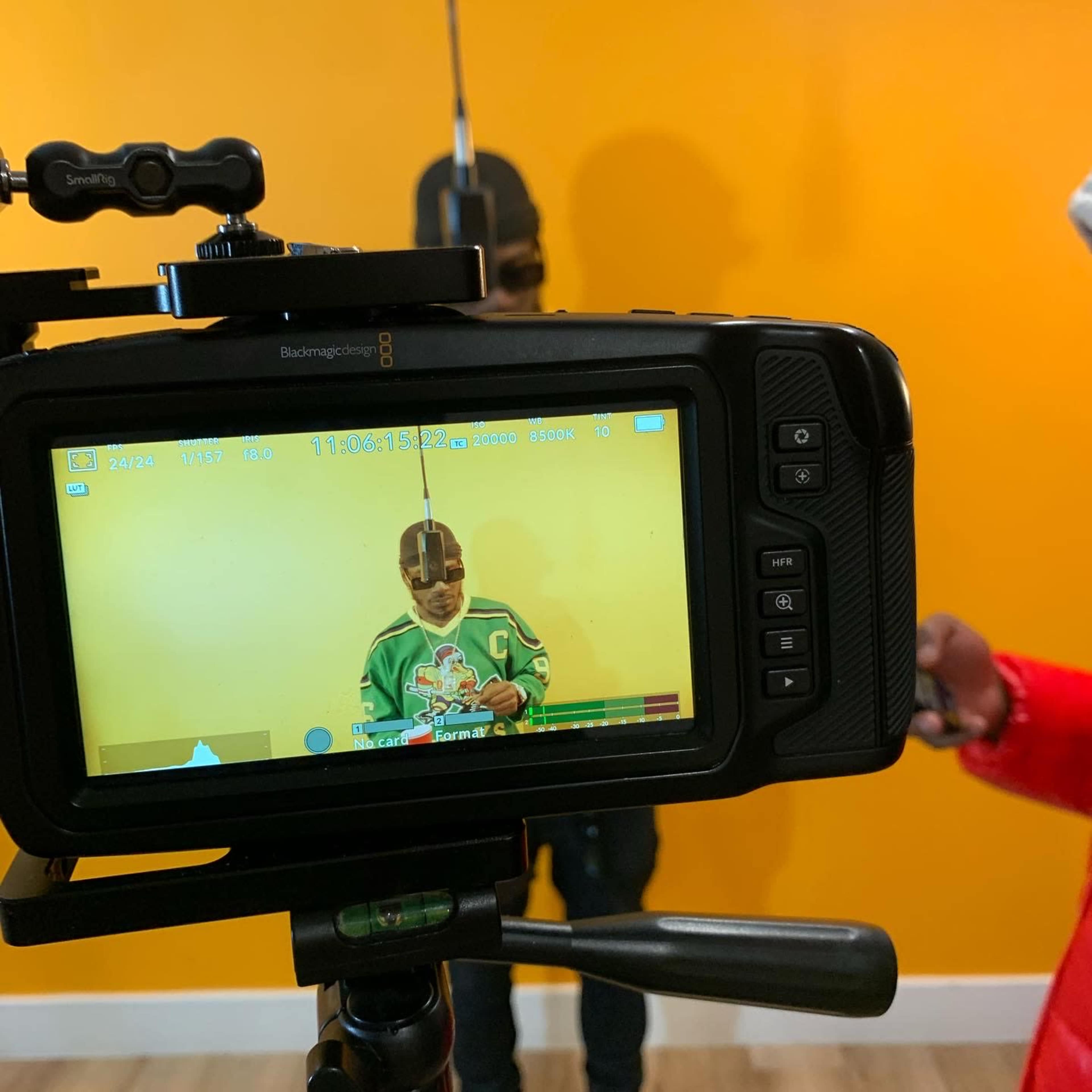 A camera on a tripod focuses on a person standing in front of a yellow wall while filming.