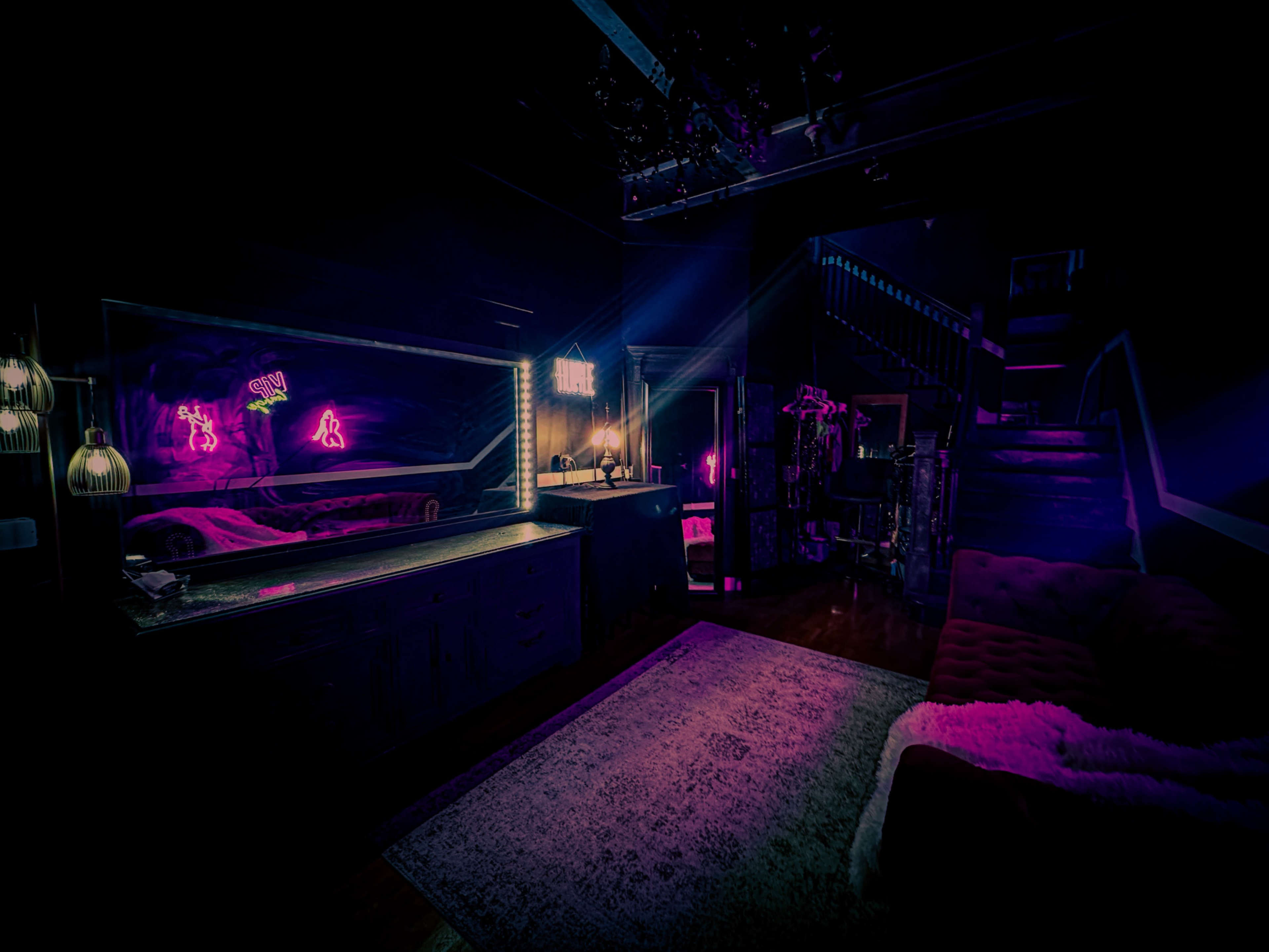 A dimly lit room features a cozy couch, a large mirror, and neon lights illuminating the space.