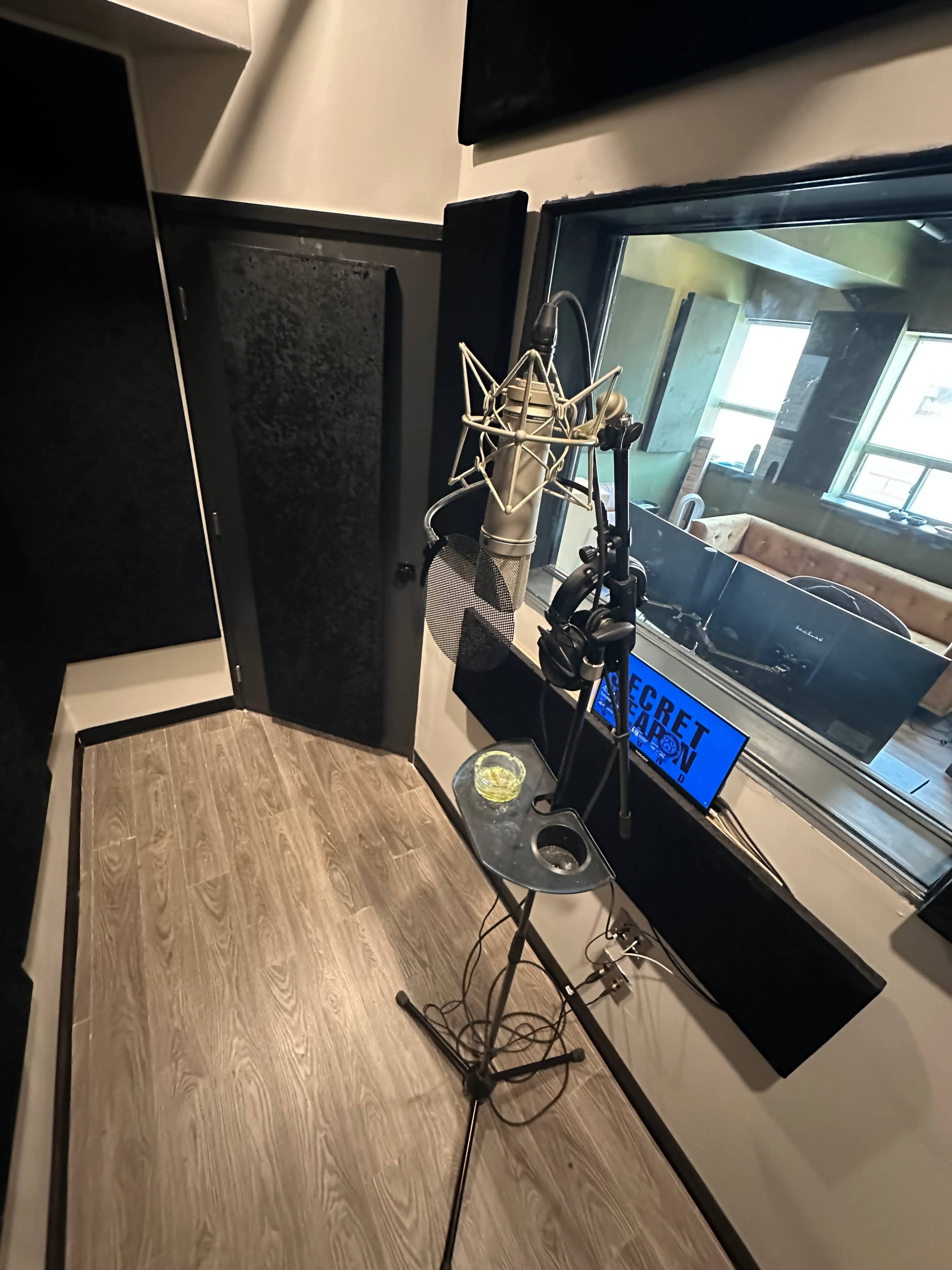 The image shows a recording booth with a microphone mounted on a stand and a small table beside it, featuring a monitor displaying the text "REGRET."