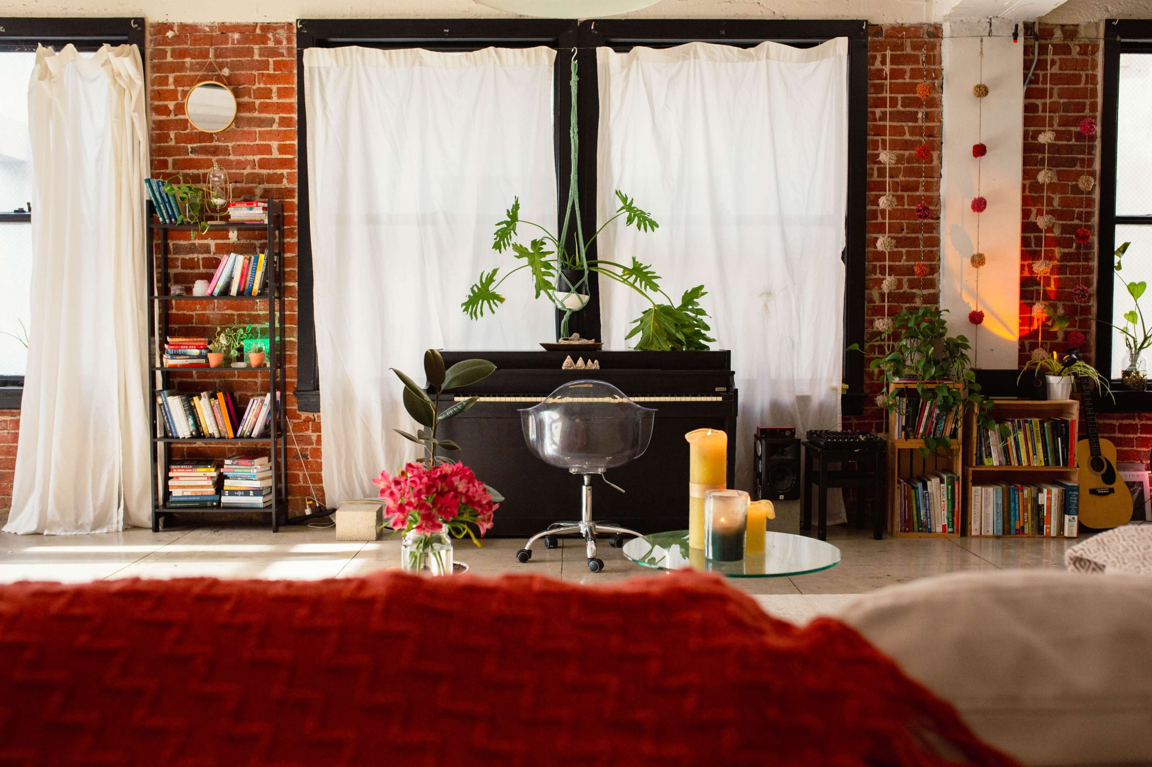A cozy living room with exposed brick walls, a black piano, bookshelves filled with books, plants, and a glass coffee table with candles and flowers.