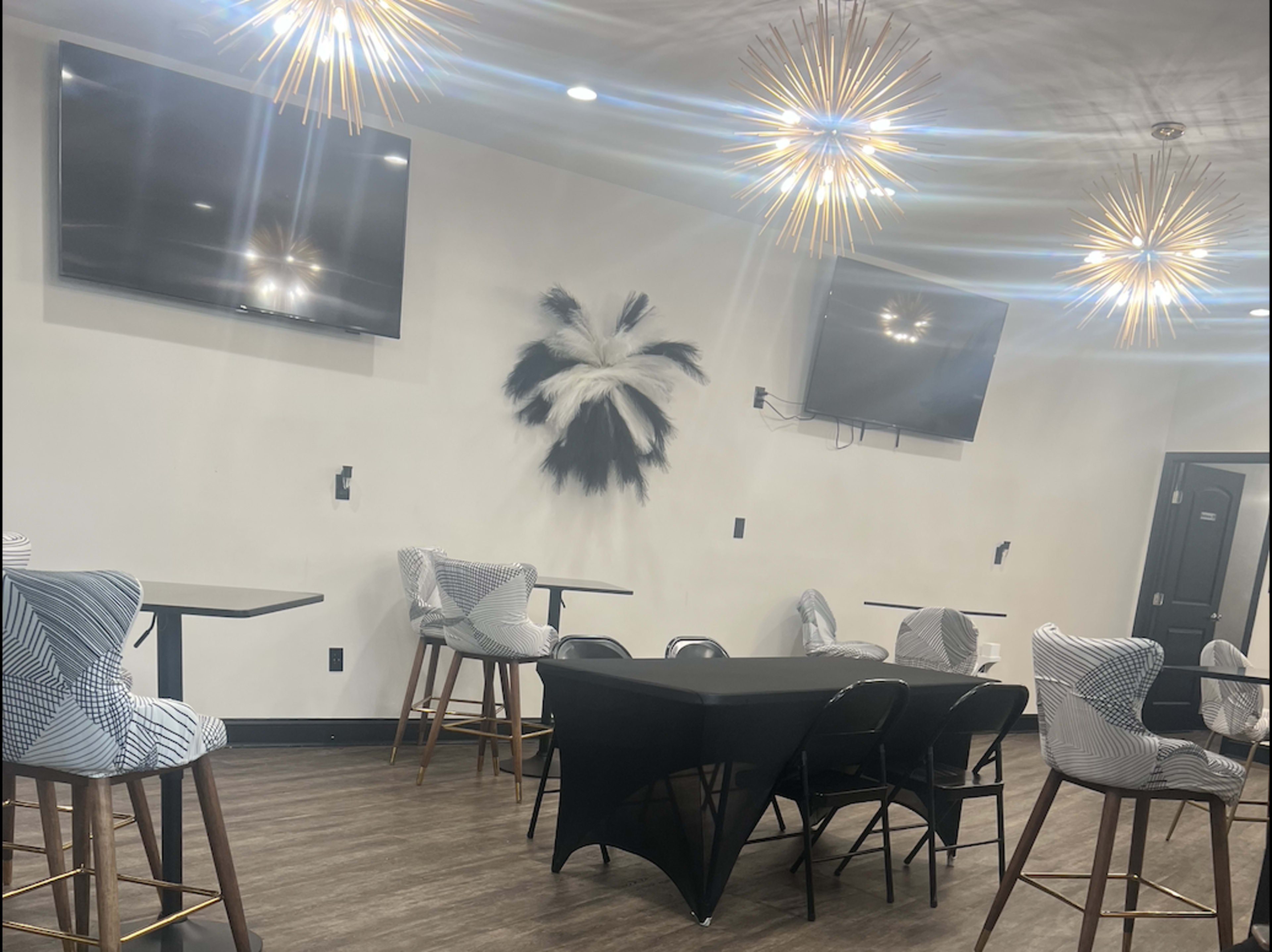 A modern room with several bar-height tables and stools, decorated with two wall-mounted TVs and a large wall art piece, along with pendant light fixtures.
